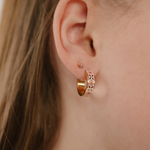 A close up of a model wearing a hoop earring made from 18k gold-plated titanium metal, with folk-art style floral patterns in pink, dark pink, and white enamel.