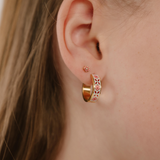 A close up of a model wearing a hoop earring made from 18k gold-plated titanium metal, with folk-art style floral patterns in pink, dark pink, and white enamel.