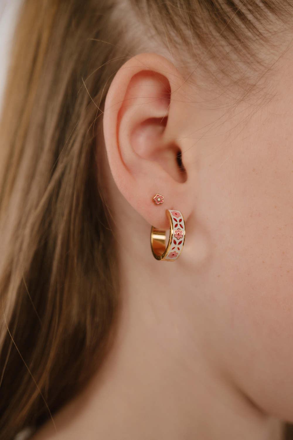 A close up of a model wearing a hoop earring made from 18k gold-plated titanium metal, with folk-art style floral patterns in pink, dark pink, and white enamel.