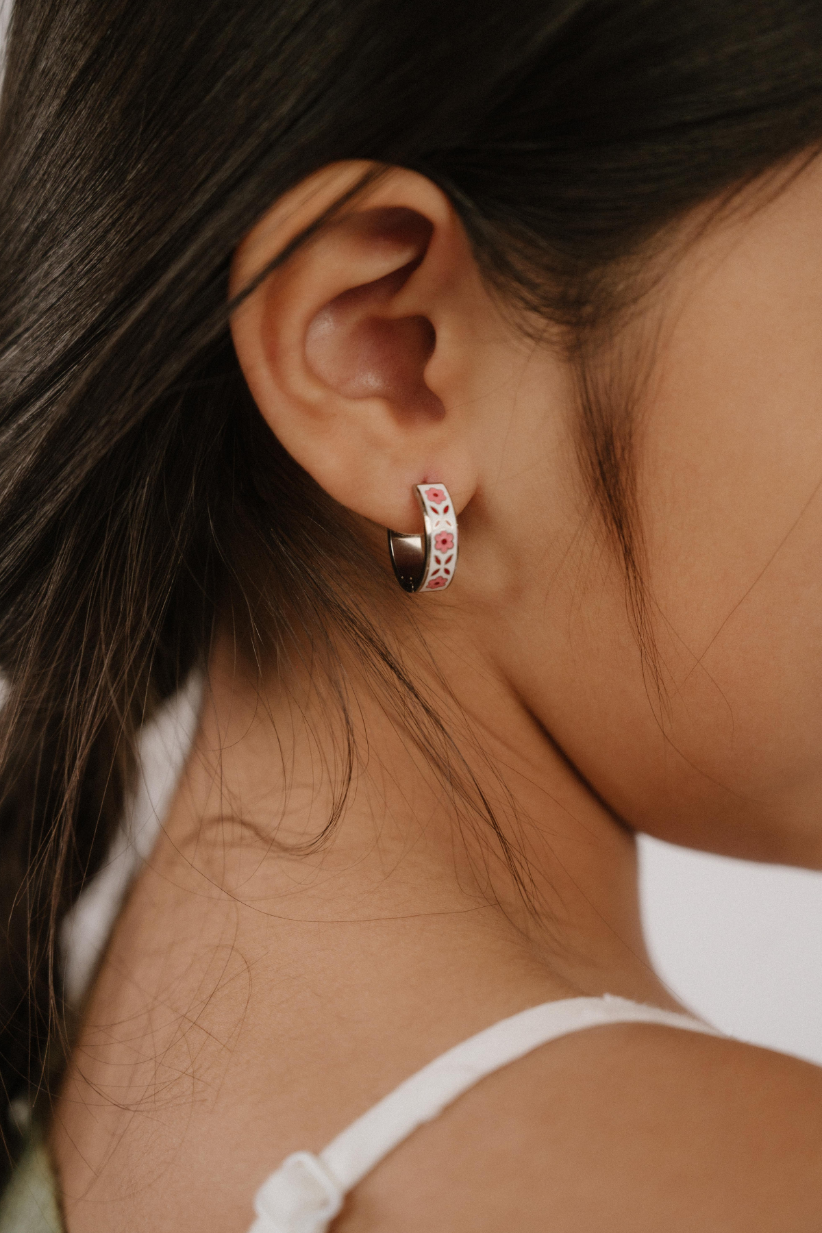 A close up of a model wearing a hoop earring made from titanium metal, with folk-art style floral patterns in pink, dark pink, and white enamel.