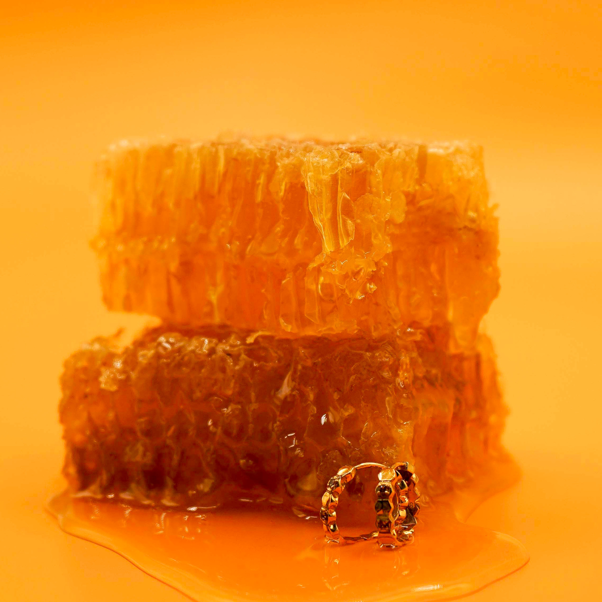 Close up of a model wearing a hoop earring made from stacked hexagon shapes with a honey colored zirconia stone set into every other hexagon shape. Hoop made from 18k gold-plated 316L Surgical Steel, ,with a honey-colored zirconia stone, sitting next to honeycomb and in a pool of honey, against a warm yellow-orange background