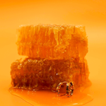 Close up of a model wearing a hoop earring made from stacked hexagon shapes with a honey colored zirconia stone set into every other hexagon shape. Hoop made from 18k gold-plated 316L Surgical Steel, ,with a honey-colored zirconia stone, sitting next to honeycomb and in a pool of honey, against a warm yellow-orange background