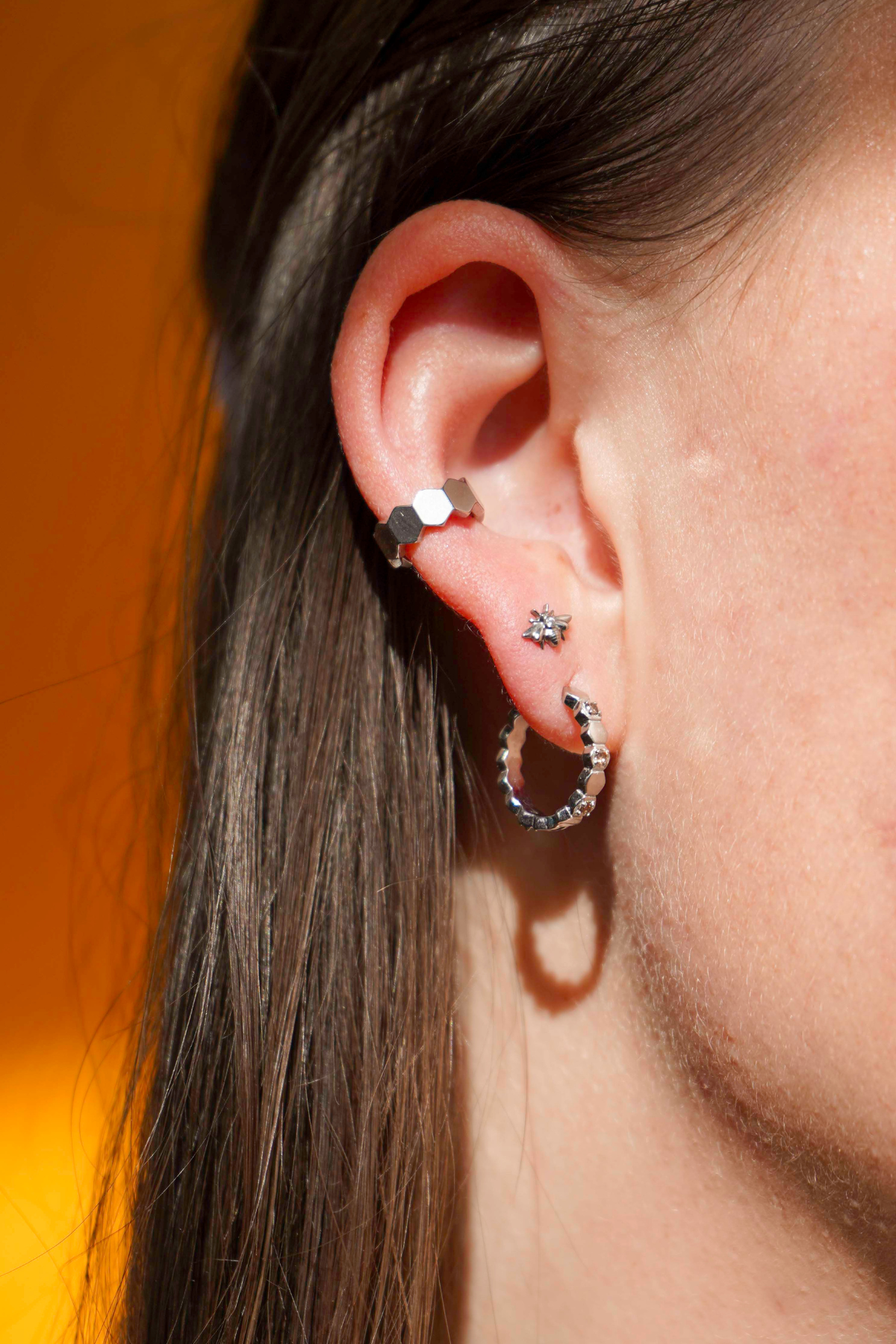 Close up of a model wearing a hoop earring made from stacked hexagon shapes with a honey colored zirconia stone set into every other hexagon shape. Hoop made from 316L Surgical Steel, with a honey-colored zirconia stone, against warm yellow background.
