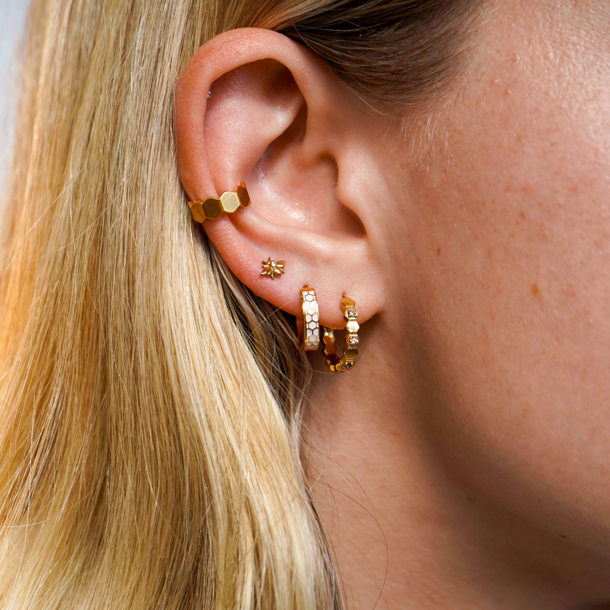 Close up of a model wearing a hoop earring made from stacked hexagon shapes with a honey colored zirconia stone set into every other hexagon shape. Hoop made from 18k gold-plated 316L Surgical Steel, , with a honey-colored zirconia stone, against a blurred background.