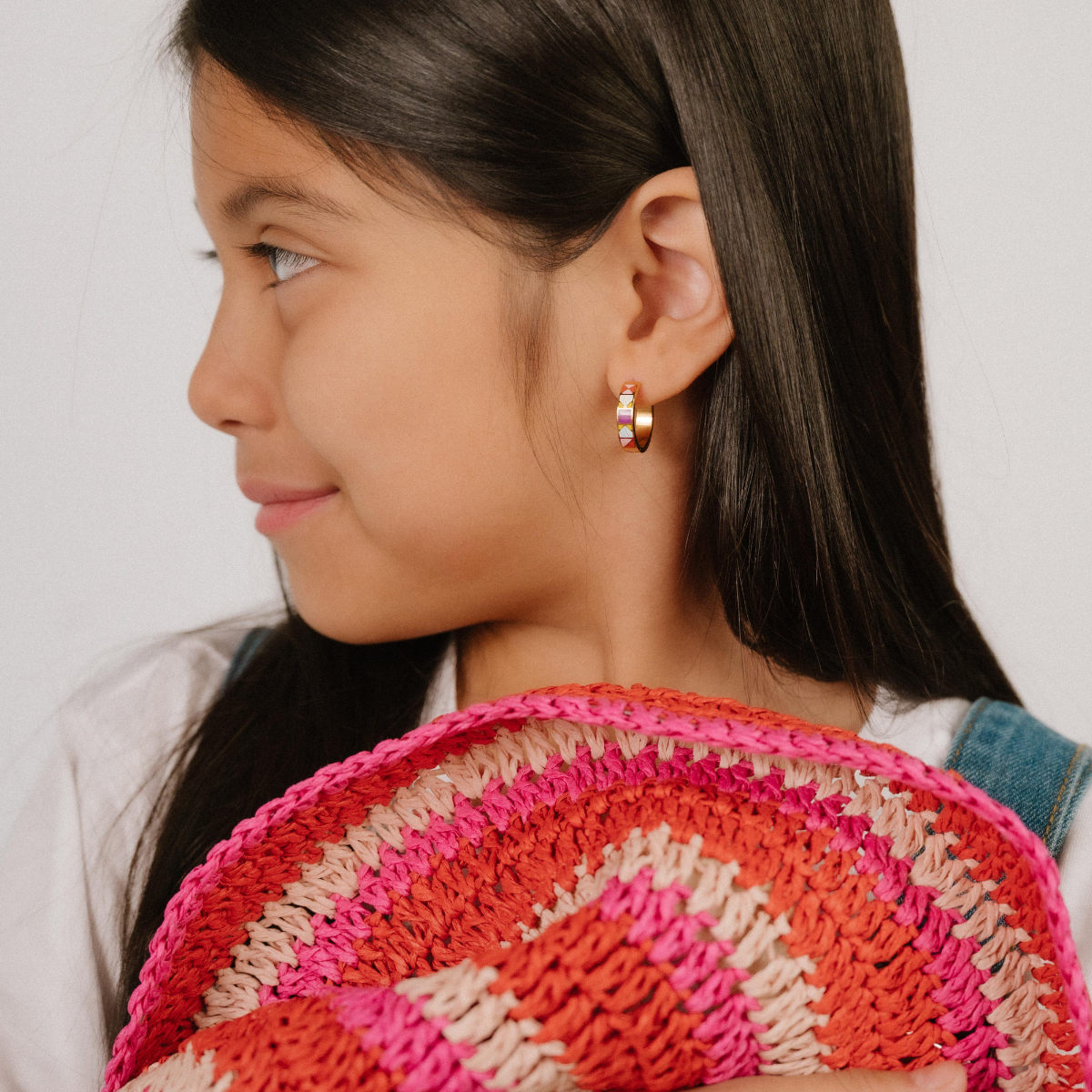 A  model wearing a hoop earring made from 18k gold-plated titanium metal, with geometric colored patterns that are reminiscent of quilt blocks in pinks, purple, blue, and green on a white background.