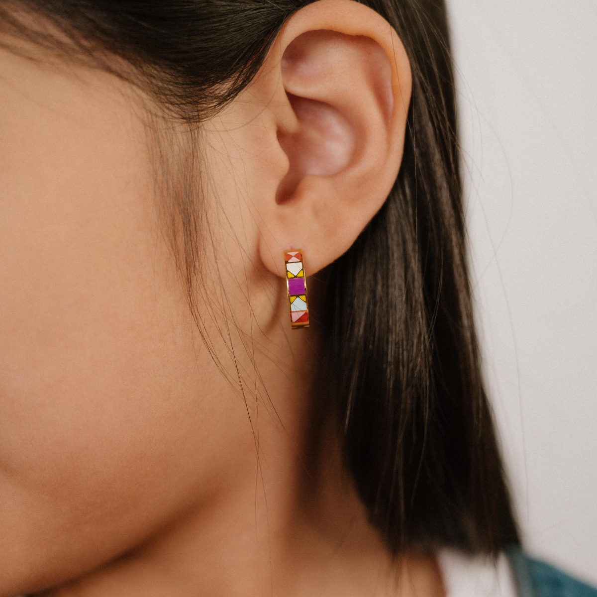 A  close up of a model wearing a hoop earring made from 18k gold-plated titanium metal, with geometric colored patterns that are reminiscent of quilt blocks in pinks, purple, blue, and green on a white background.