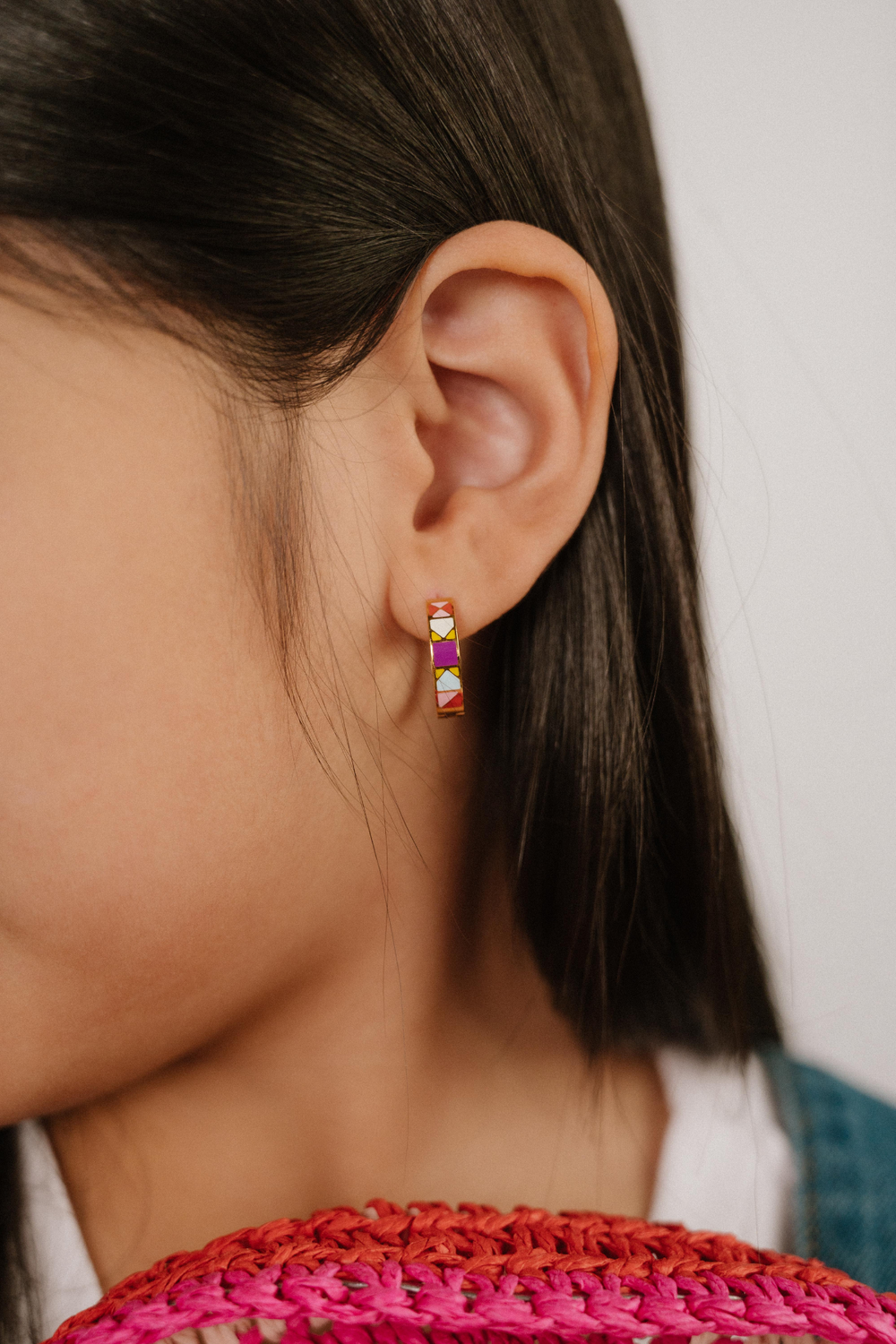 A  close up of a model wearing a hoop earring made from 18k gold-plated titanium metal, with geometric colored patterns that are reminiscent of quilt blocks in pinks, purple, blue, and green on a white background.