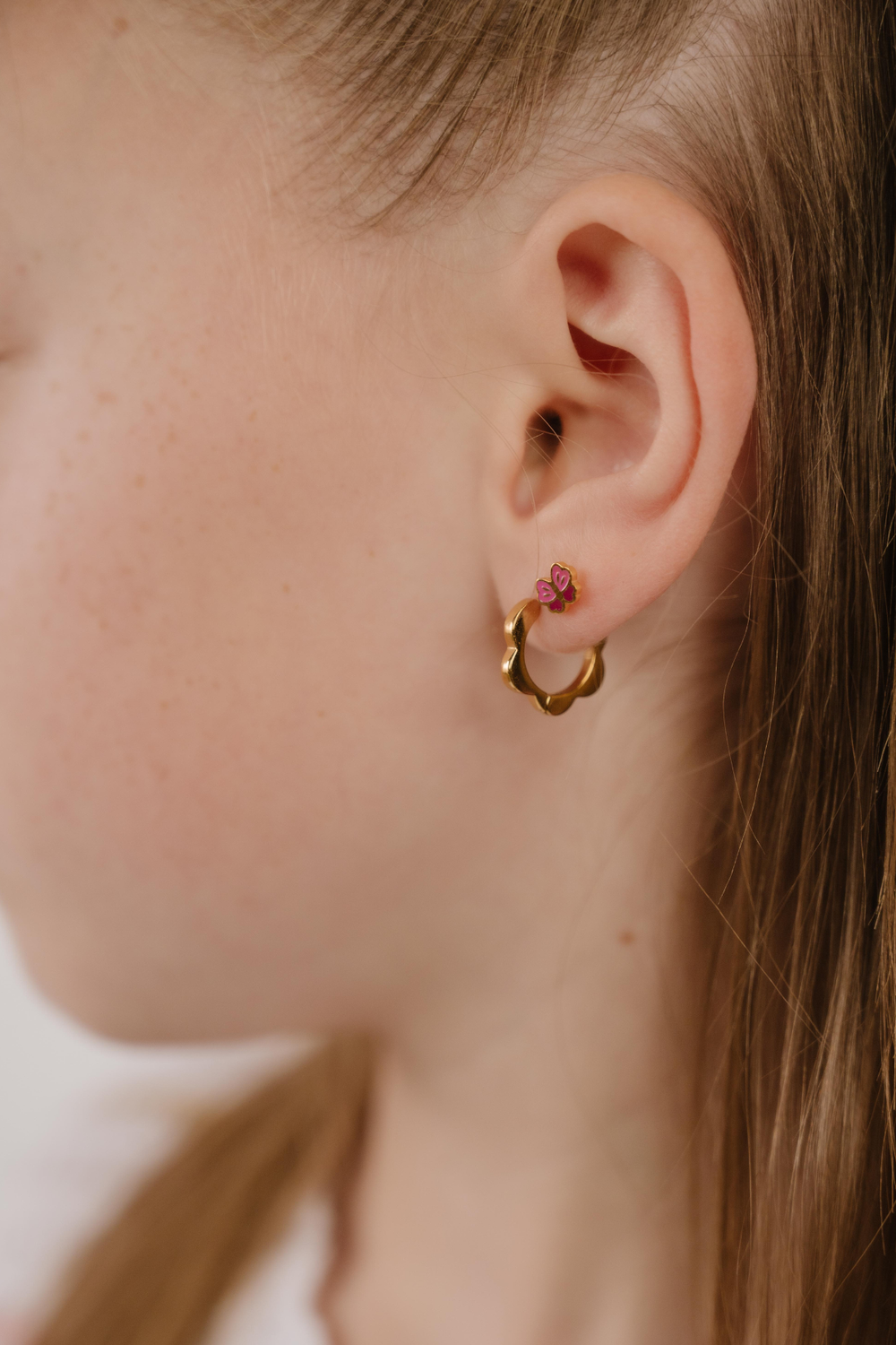 A close up of a model wearing a hoop earring with a softly scalloped edge resembling a daisy, made from 18k gold-plated titanium metal