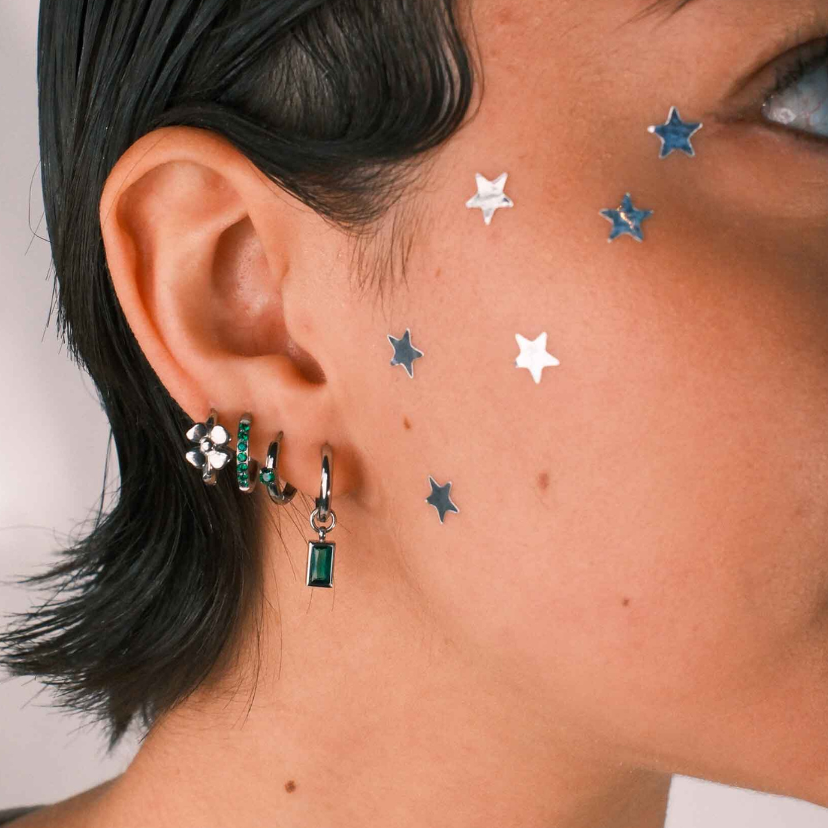 Close-up of a person wearing multiple earrings including a hoop with a channel of emerald green zirconia stones