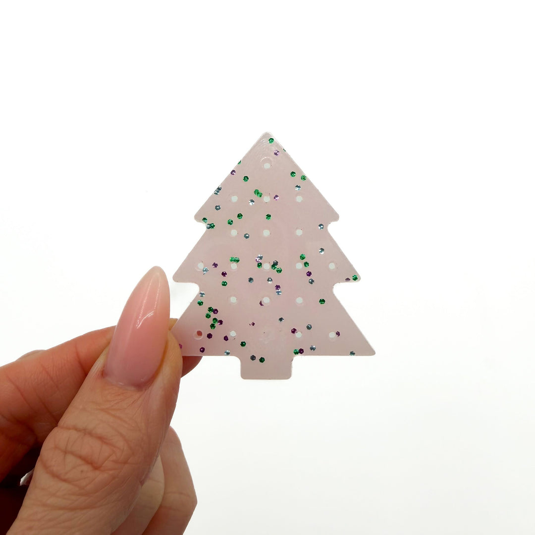 Hand holding a light pink, glittery Christmas tree-shaped earring organizer with holes in it, against a white background.
