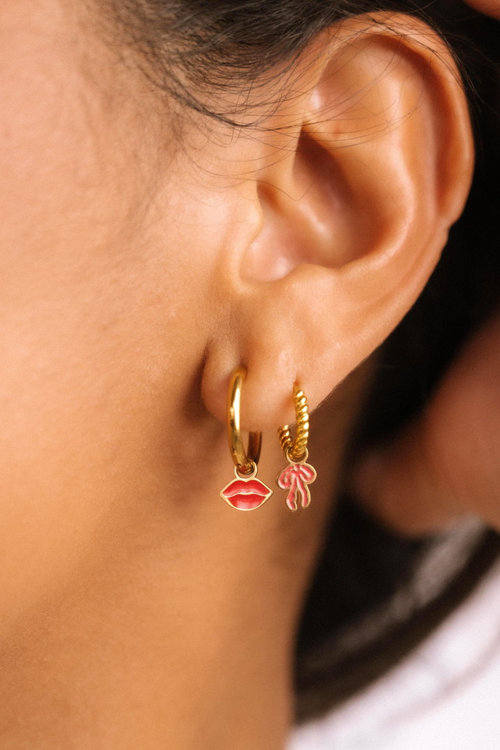 Close-up of an ear wearing gold hoop earrings with lip and bow charms.