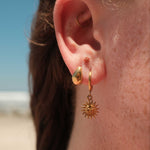Close-up of an ear wearing gold hoop earrings with a sun design, with blurred background.