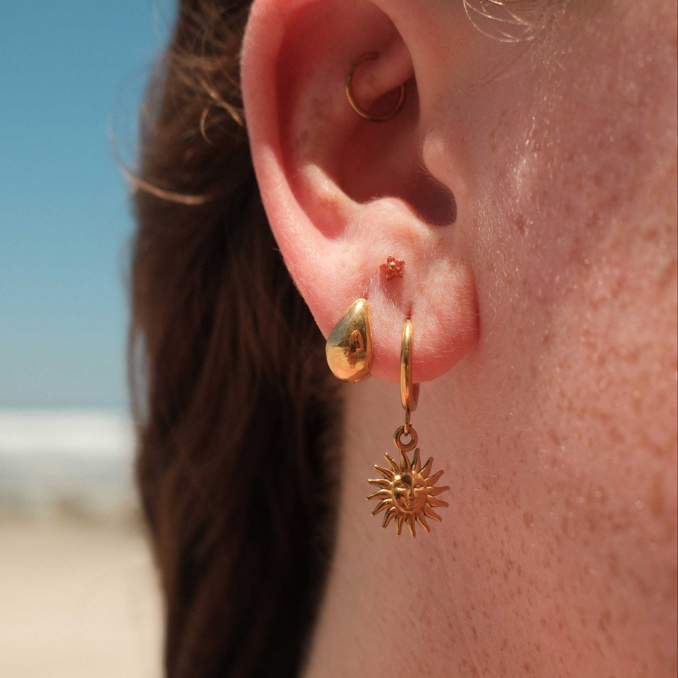 Close-up of an ear wearing gold hoop earrings with a sun design, with blurred background.