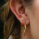 Close-up of an ear with gold hoop earrings and orange studs against a blurred background.