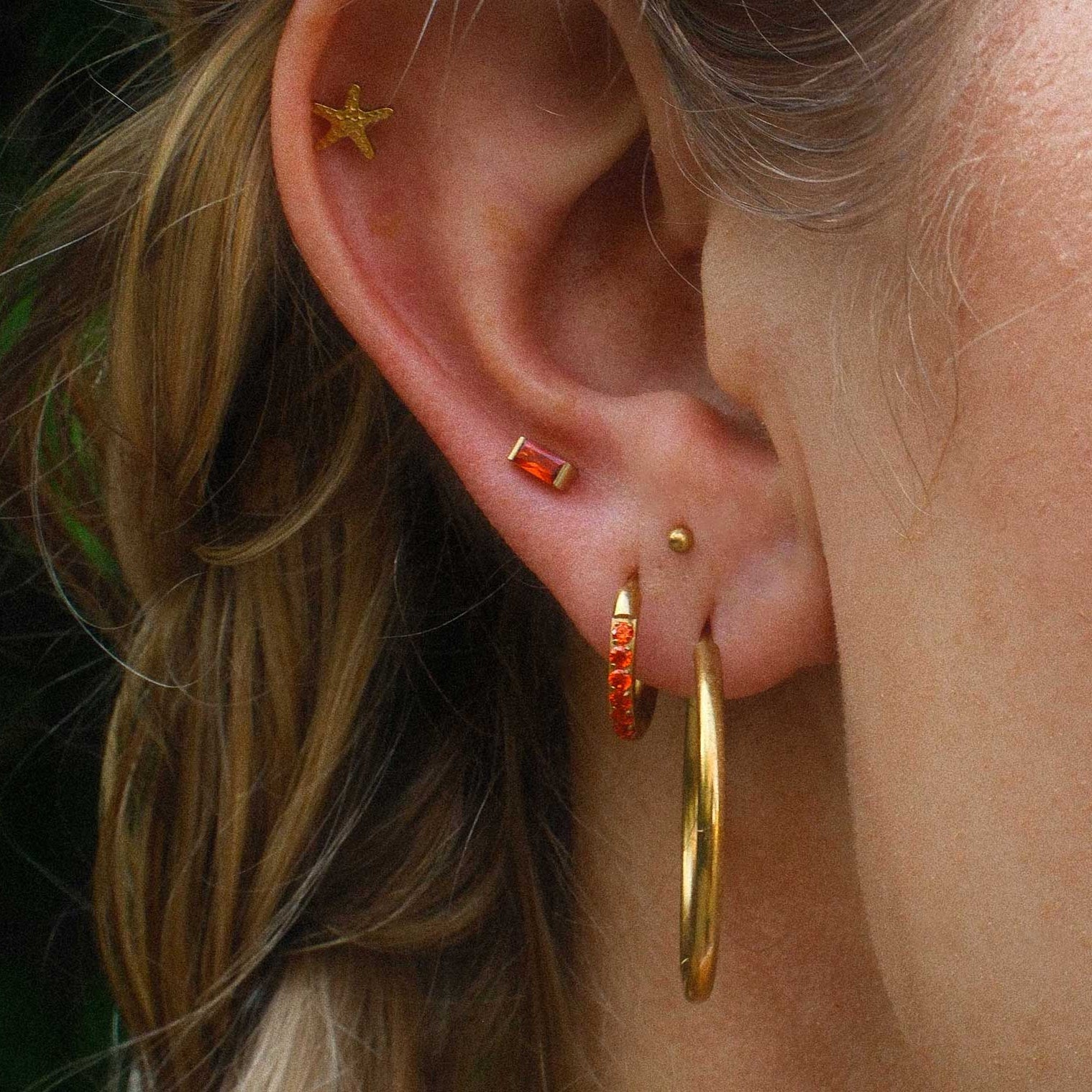 Close-up of an ear with gold hoop earrings and orange studs against a blurred background.