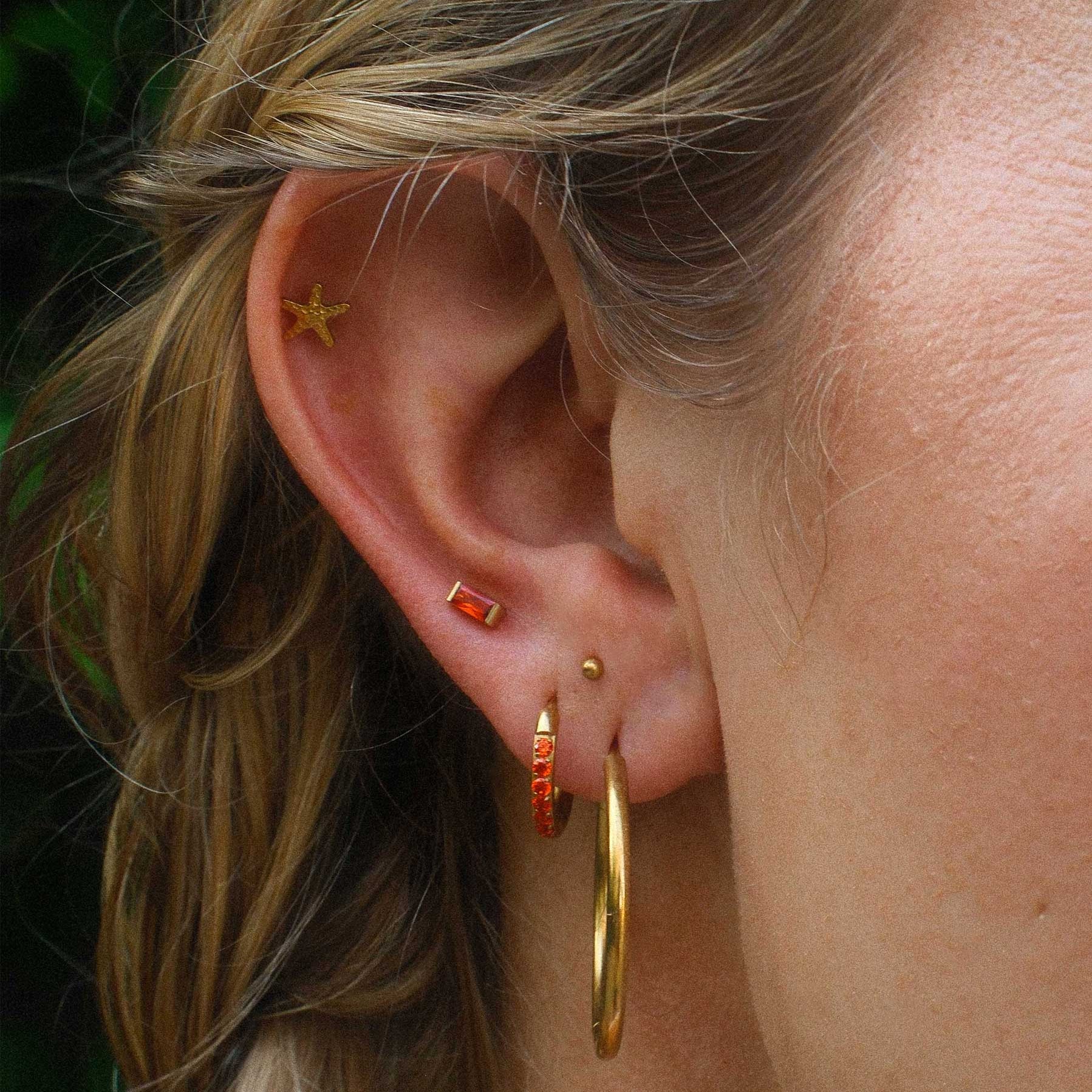 Close-up of an ear with gold hoop earrings and orange studs against a blurred background.
