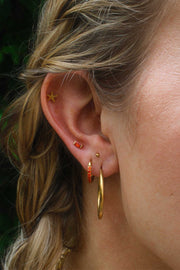Close-up of an ear with gold hoop earrings and orange studs against a blurred background.