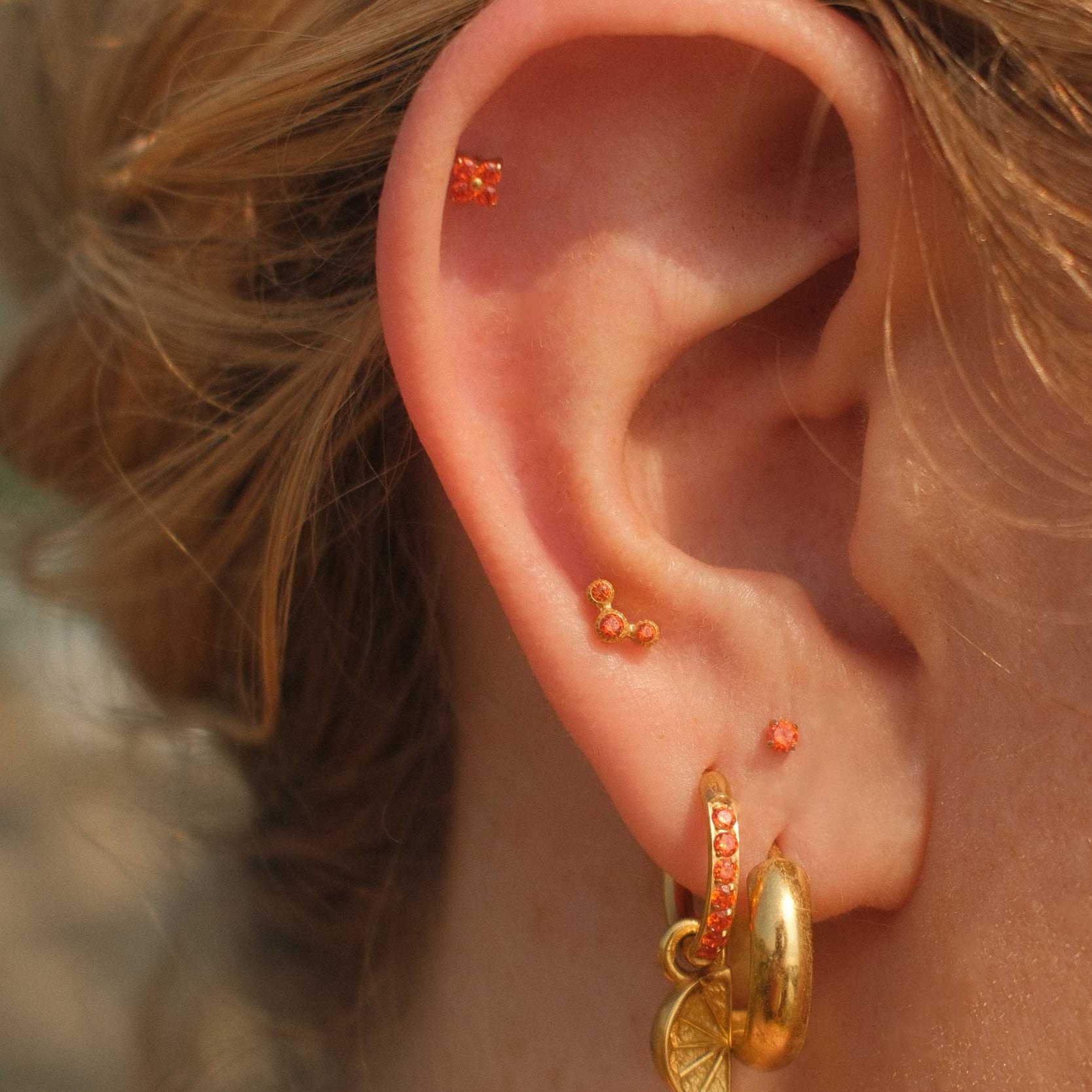 Close-up of an ear with gold earrings and small orange studs against a blurred natural background