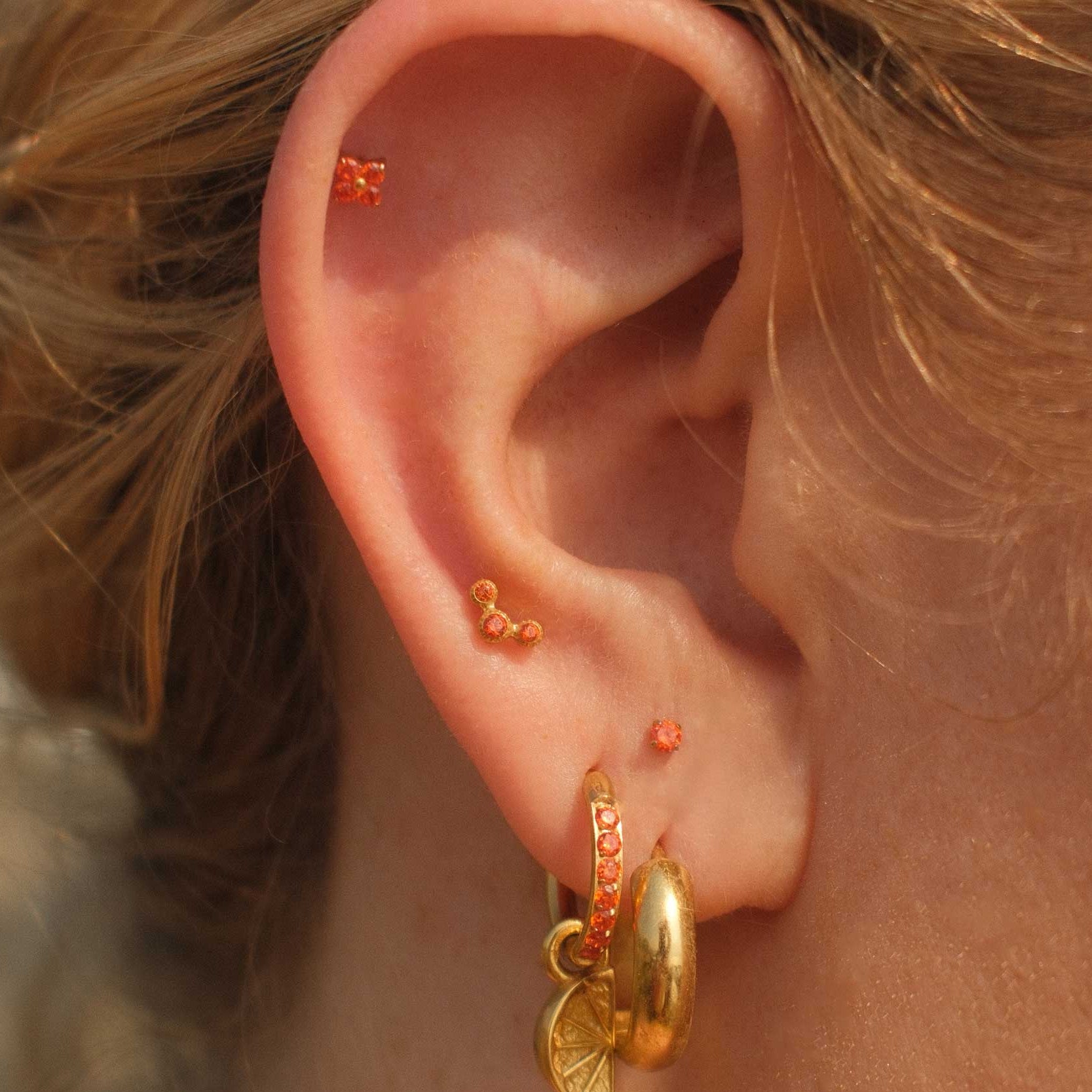 Close-up of an ear with gold earrings and small orange studs against a blurred natural background