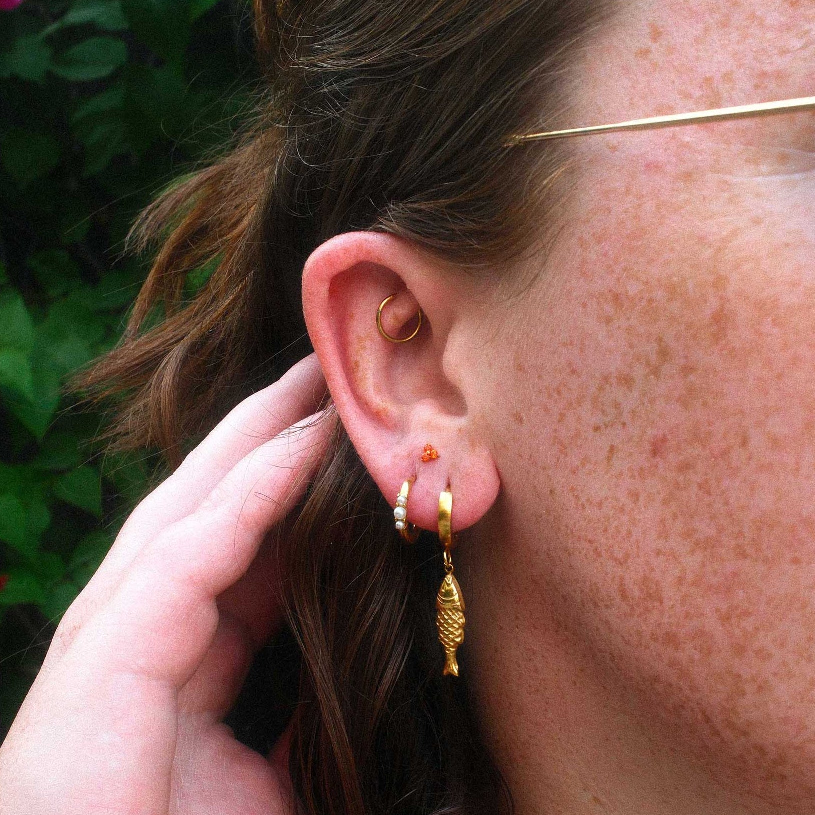 Close-up of a person wearing gold hoop earrings and an orange flat back stud with a blurred background