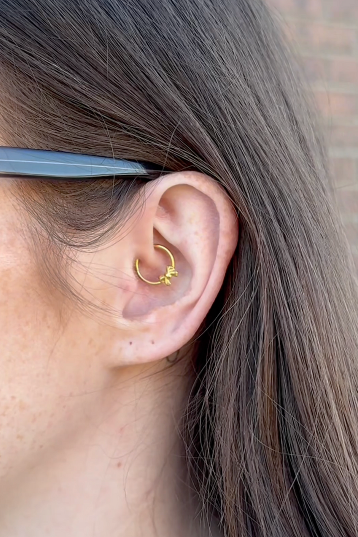 Close-up of an ear with a gold bow daith hoop earring, hair partially covering the face.