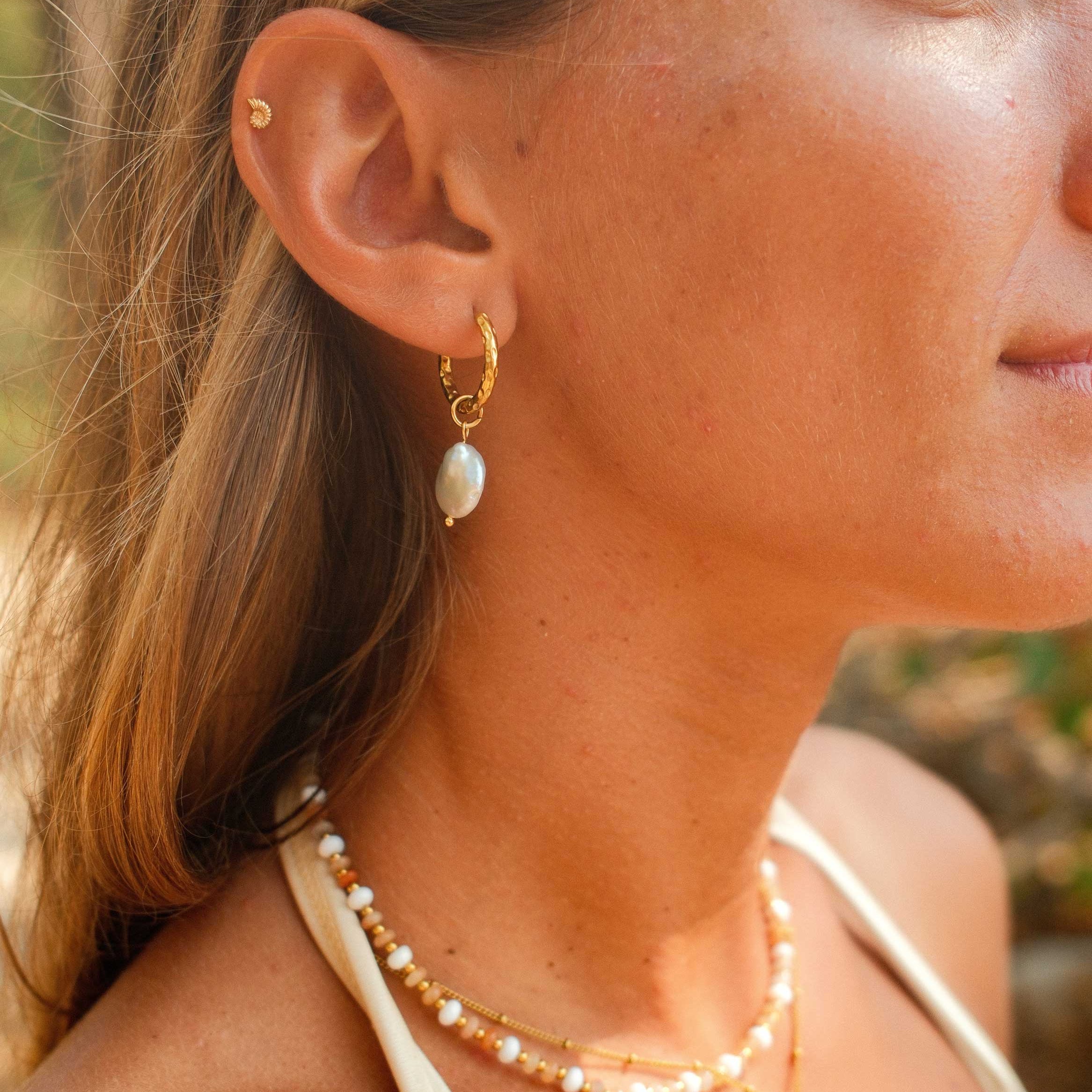 Close-up of a woman wearing gold earrings and a necklace with a blurred natural background