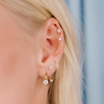 Close-up of a person wearing multiple gold earrings with clear gemstones against a blurred background.