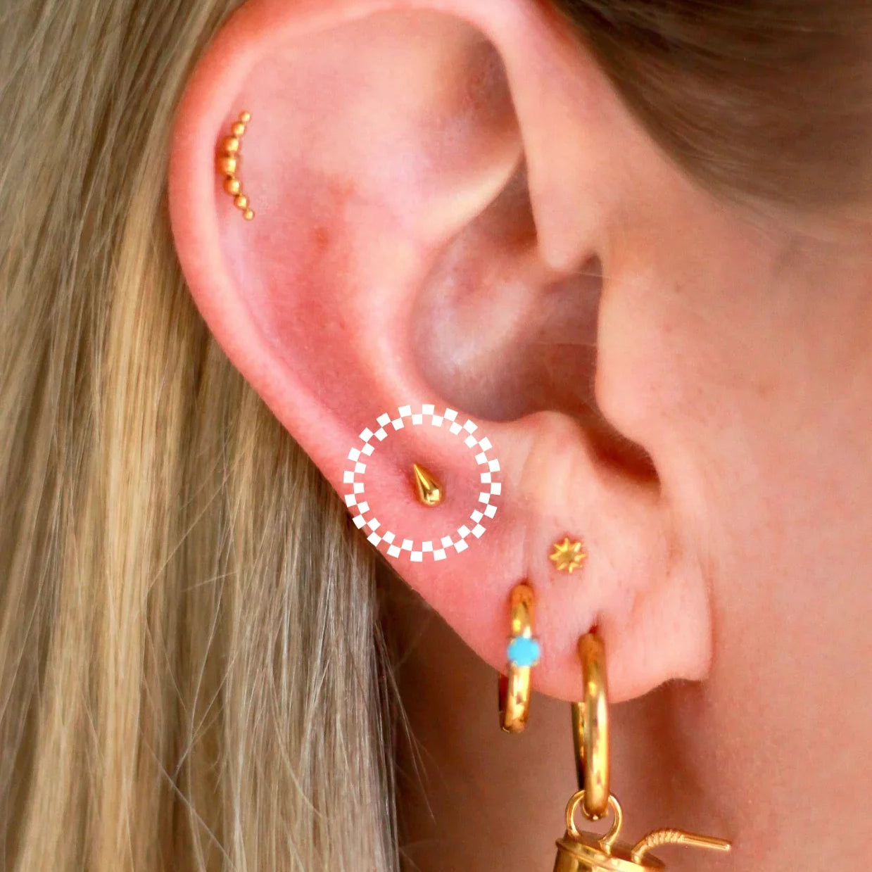 Close up of an ear with a lot of piercings, featuring gold flat backs studs and hoops. One flat back stud has been circled as the feature product in this listing. It is a gold, 3D teardrop shape.