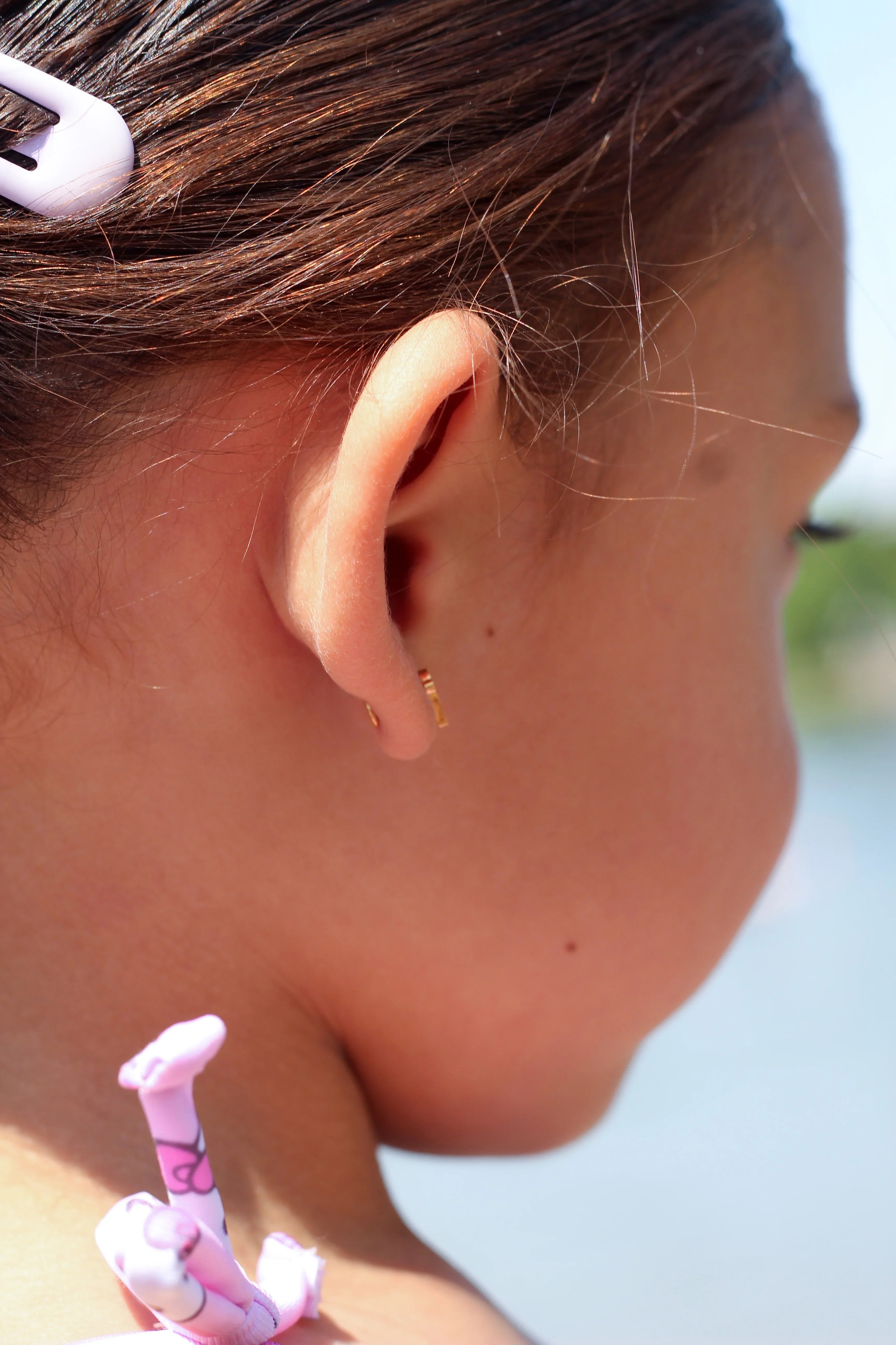 Close-up of a child's ear with a side view of a flat back earring, hair clip, and pink hair tie.