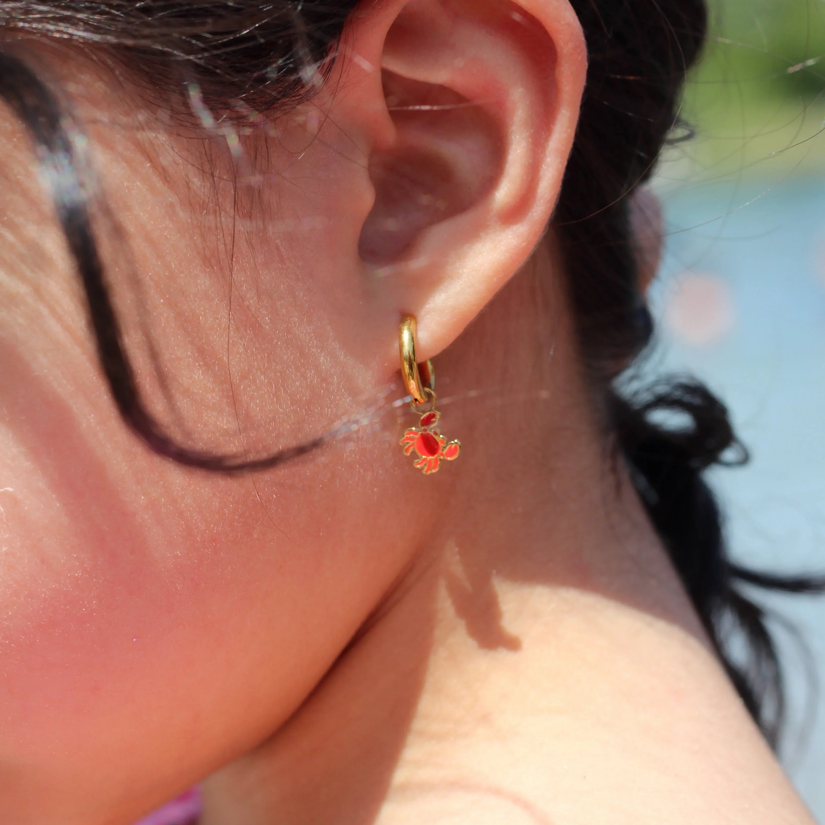 Close-up of a person's ear with a red crab charm on a gold hoop earring, outdoors.