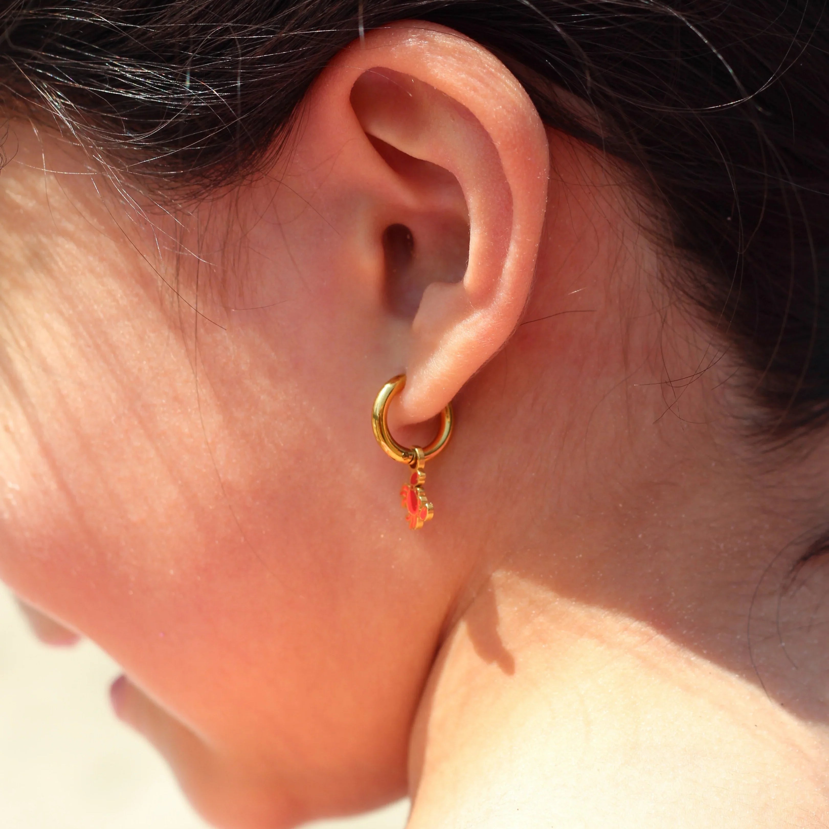 Close-up of an ear wearing a gold hoop earring with red crab charm on it.