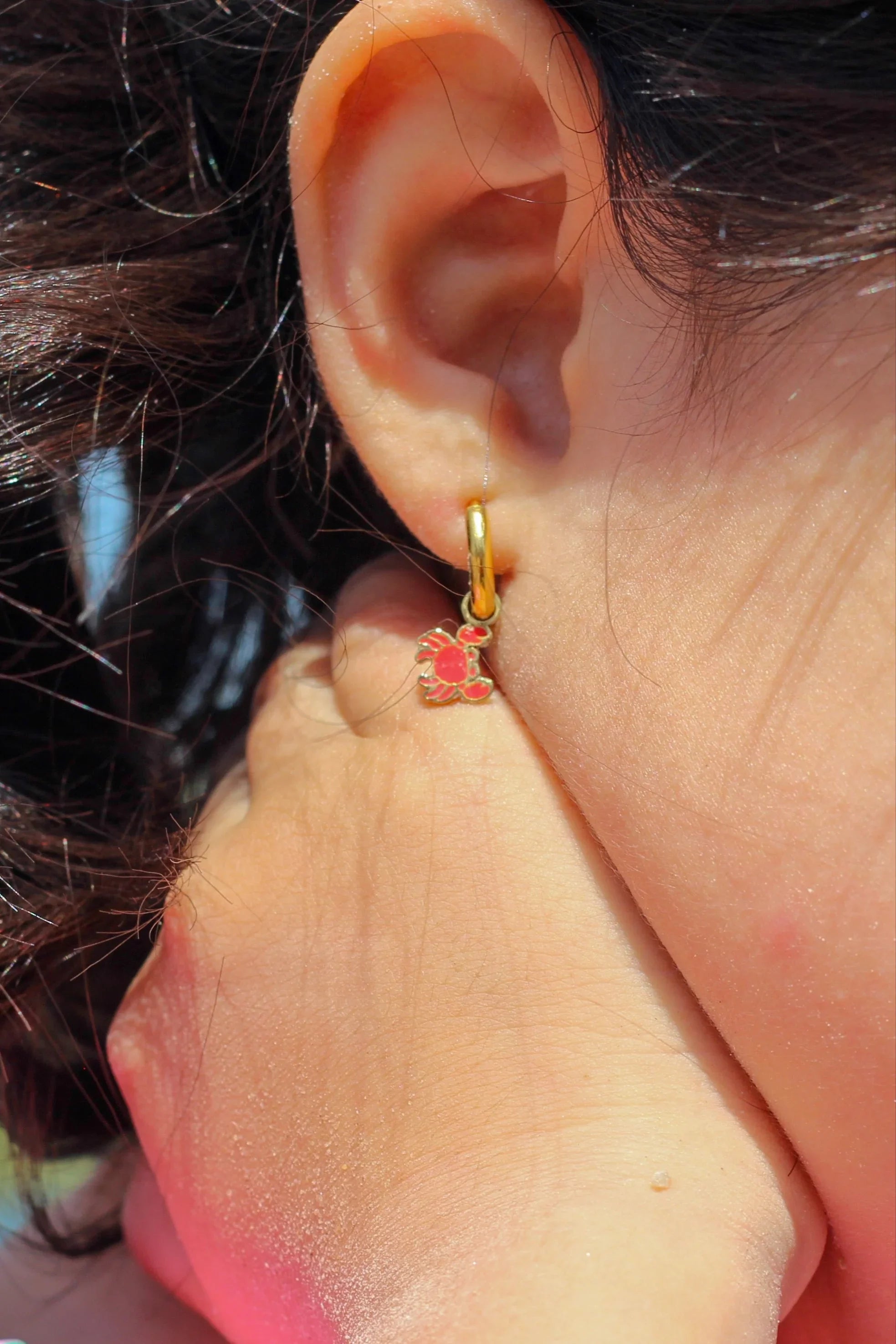 Close-up of an ear wearing a gold hoop earring with red crab charm on it.