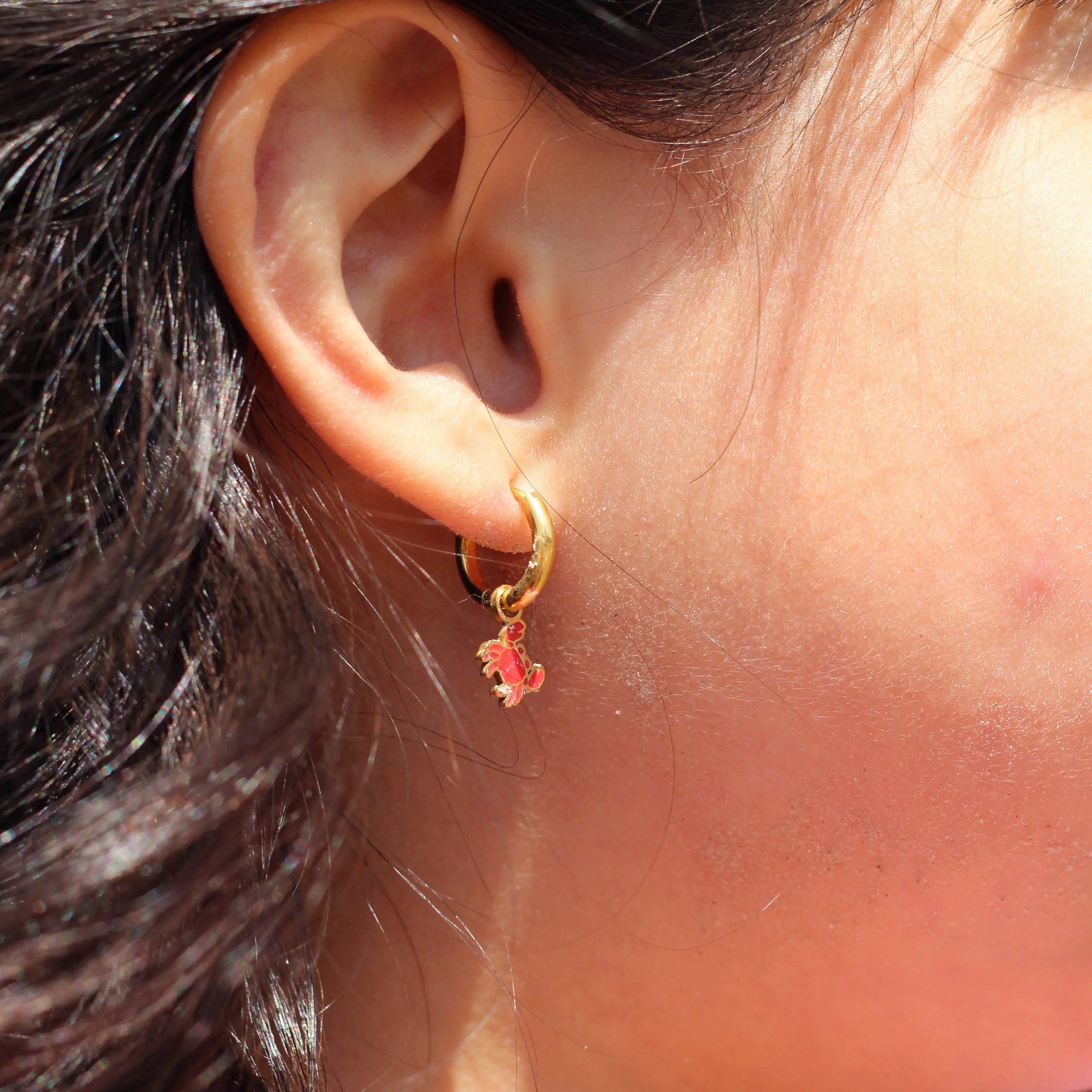Close-up of an ear wearing a gold hoop earring with red crab charm on it.