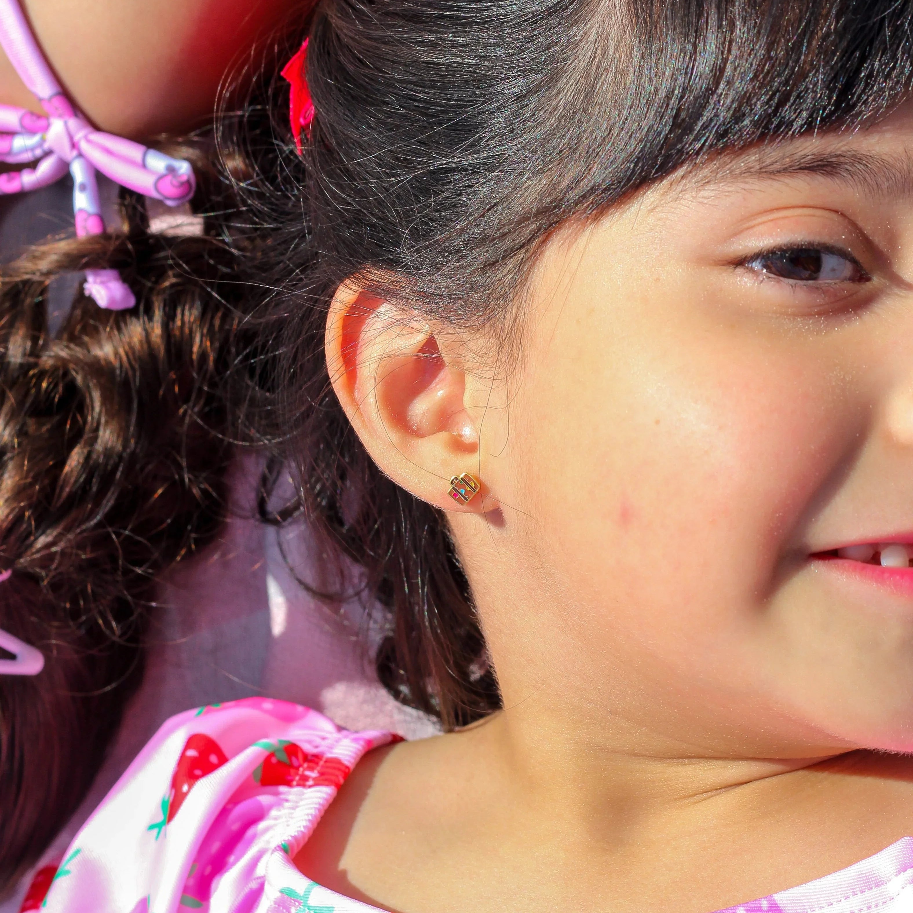Close-up of a girl wearing a brown suitcase flat back stud earring, wearing a pink swimsuit with strawberry pattern.