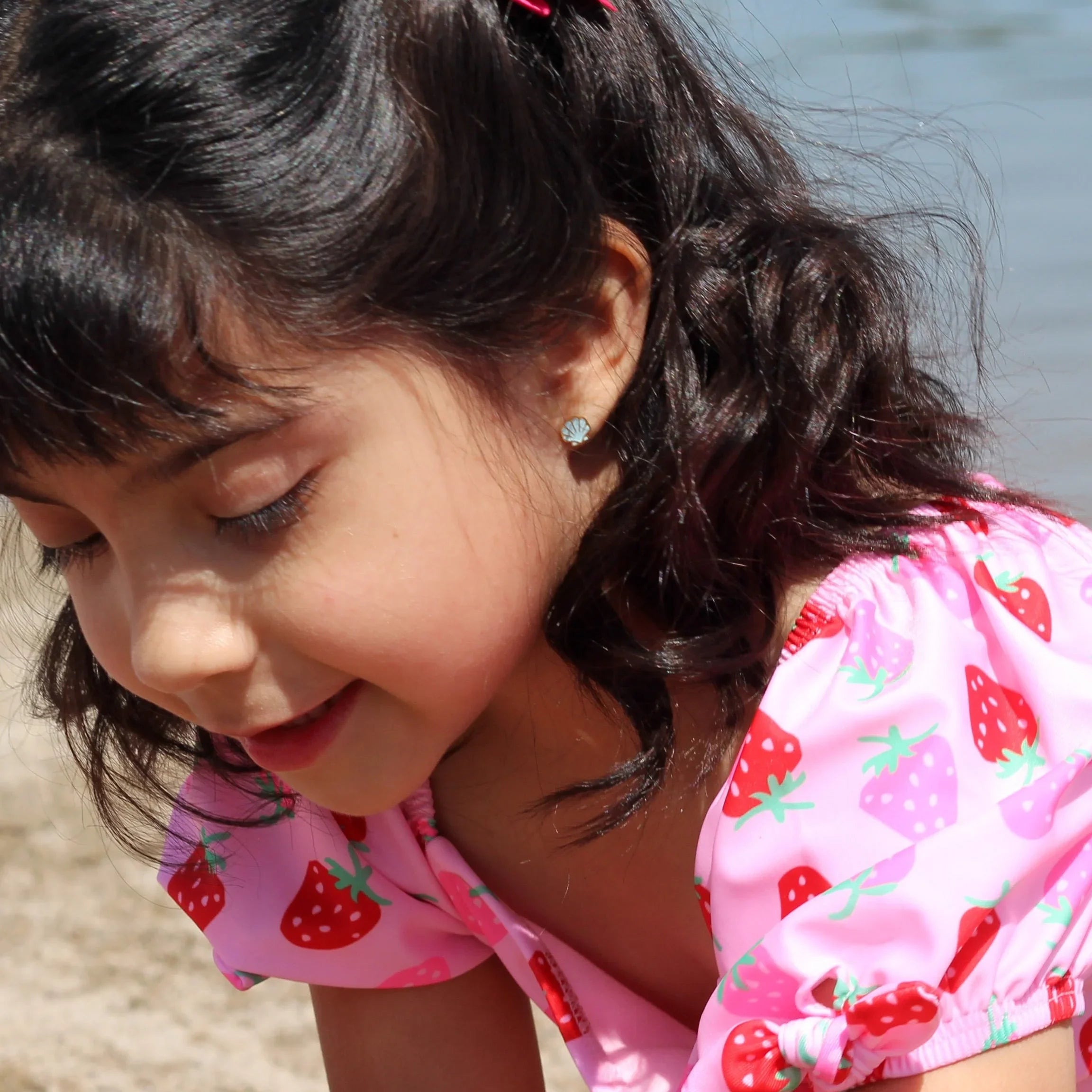 Young girl with a blue shell-shaped earring in a pink swimsuit playing by a body of water.