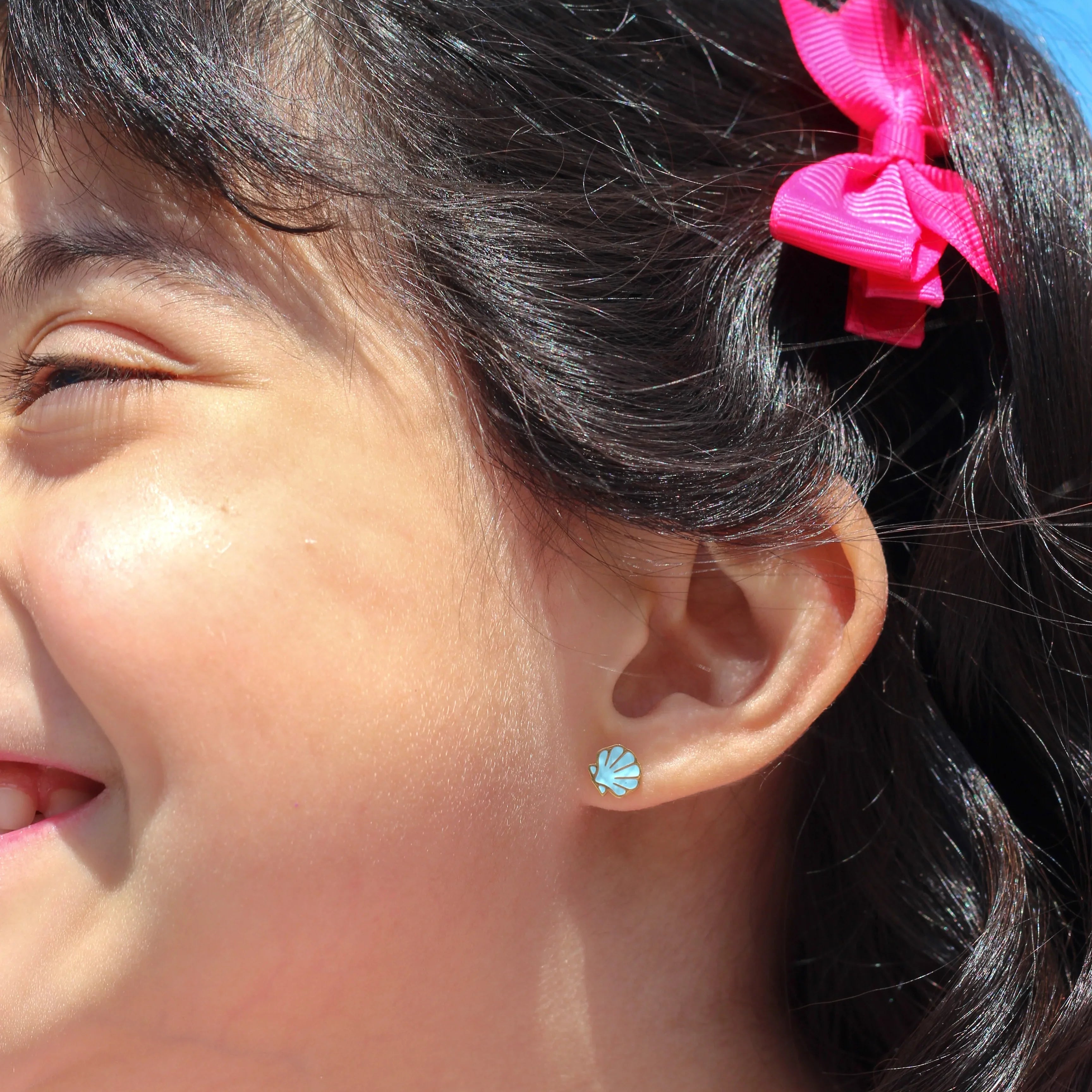 Close-up of a child with a pink hair clip and blue shell earring against a blue sky.