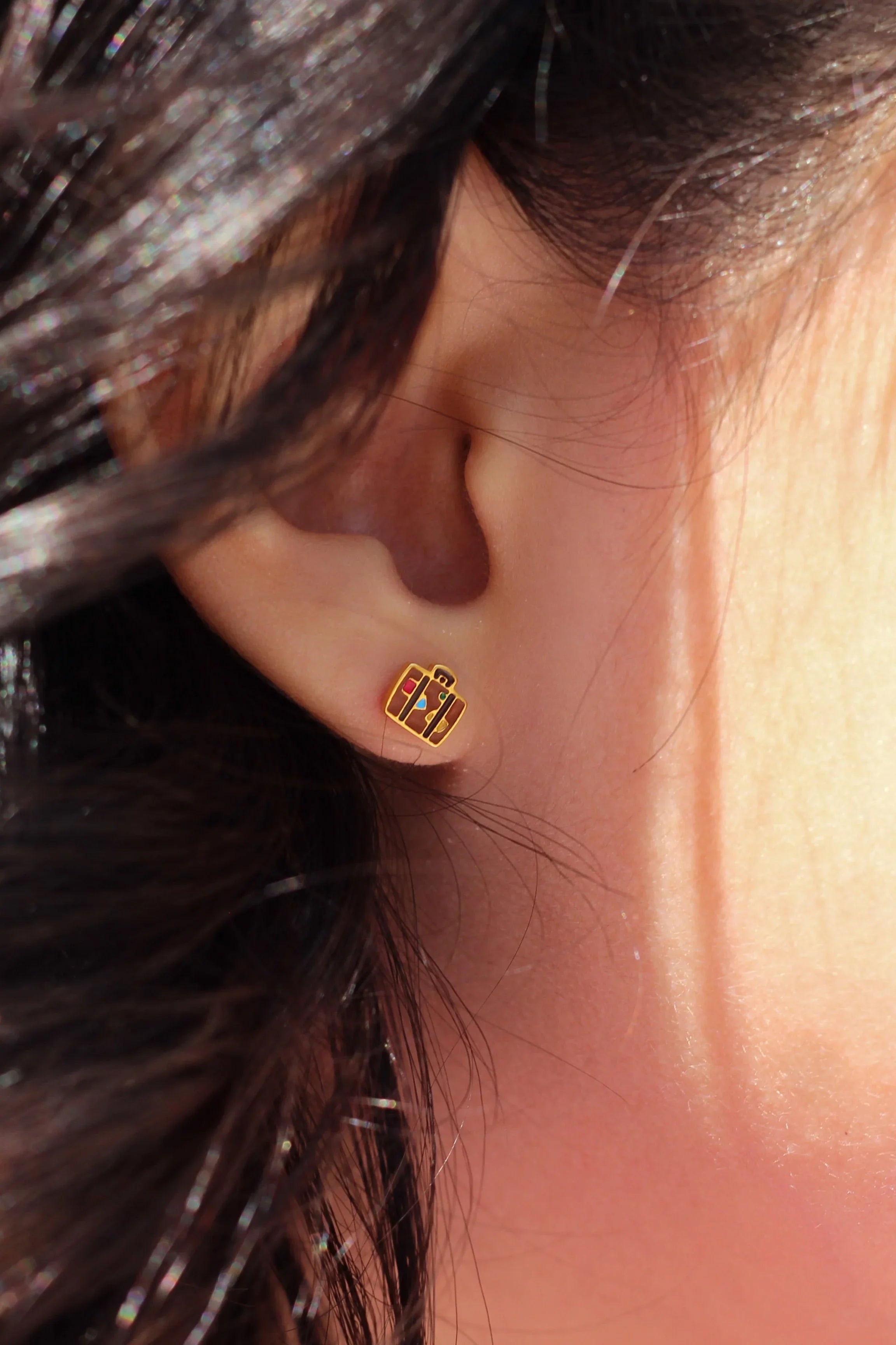 Close-up of an ear with a gold and brown suitcase-shaped earring, hair partially covering the face.