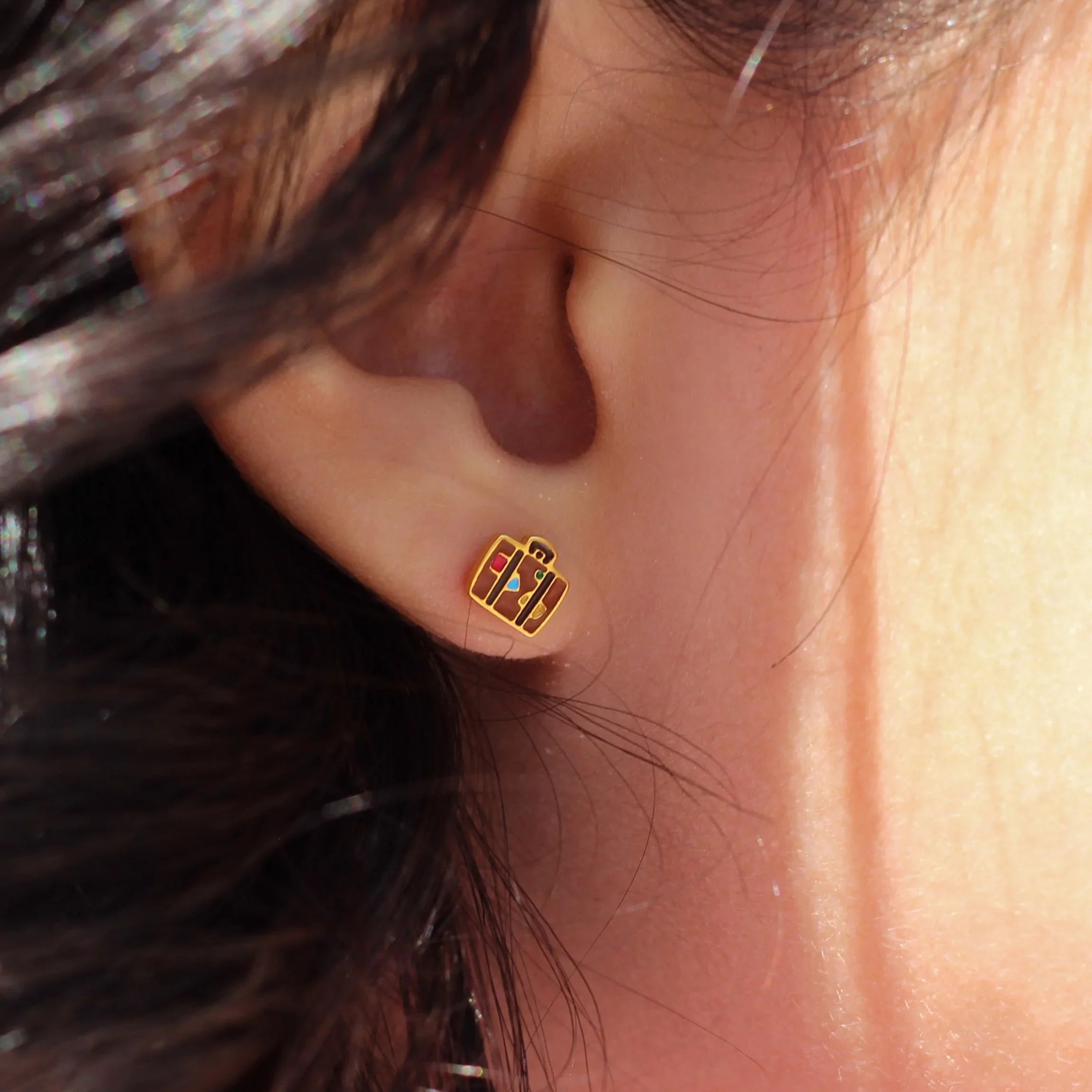 Close-up of an ear with a gold and brown suitcase-shaped earring, hair partially covering the face.
