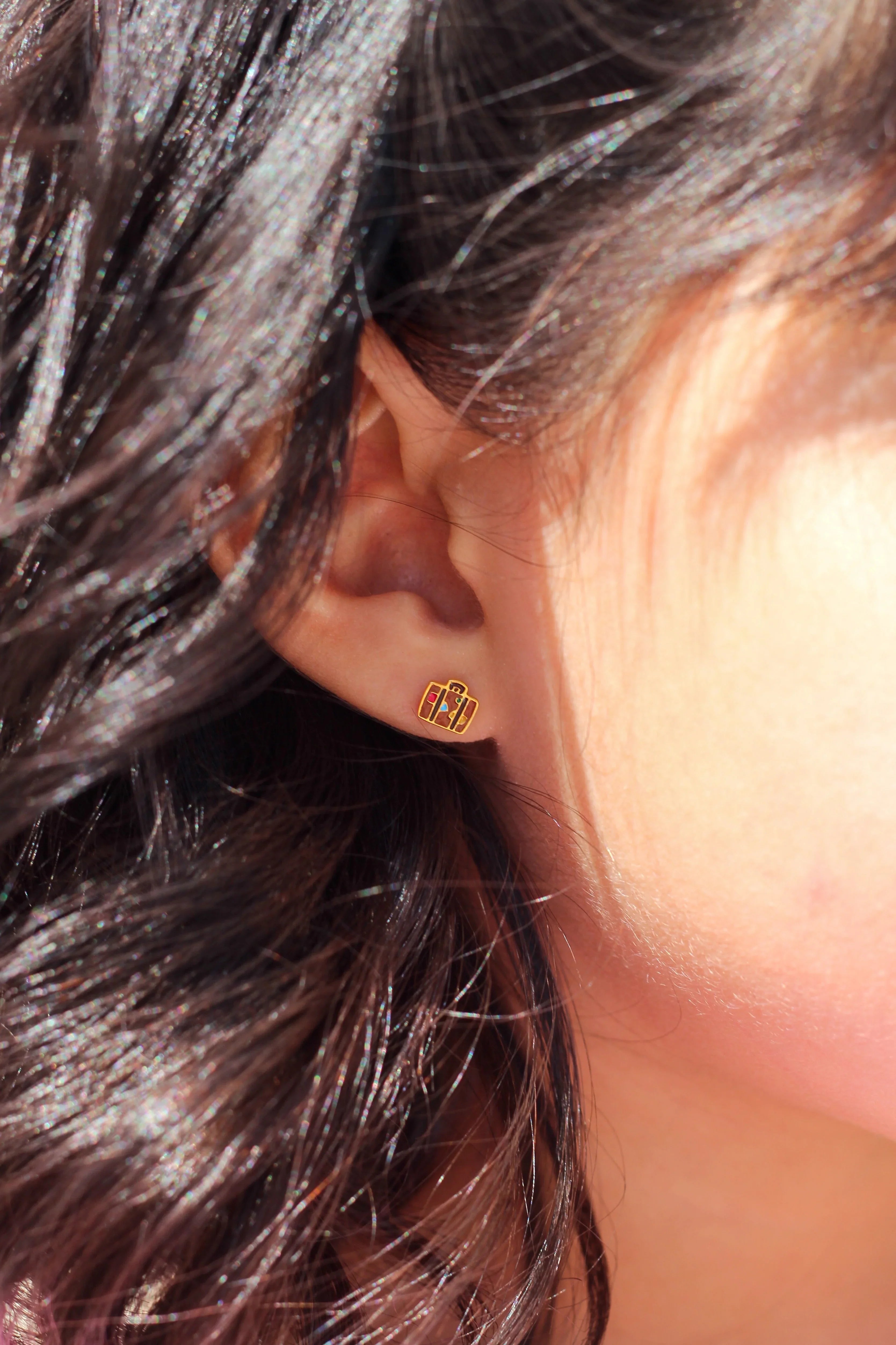 Close-up of an ear wearing a brown enamel suitcase-shaped earring with colorful shapes on it, surrounded by dark hair.