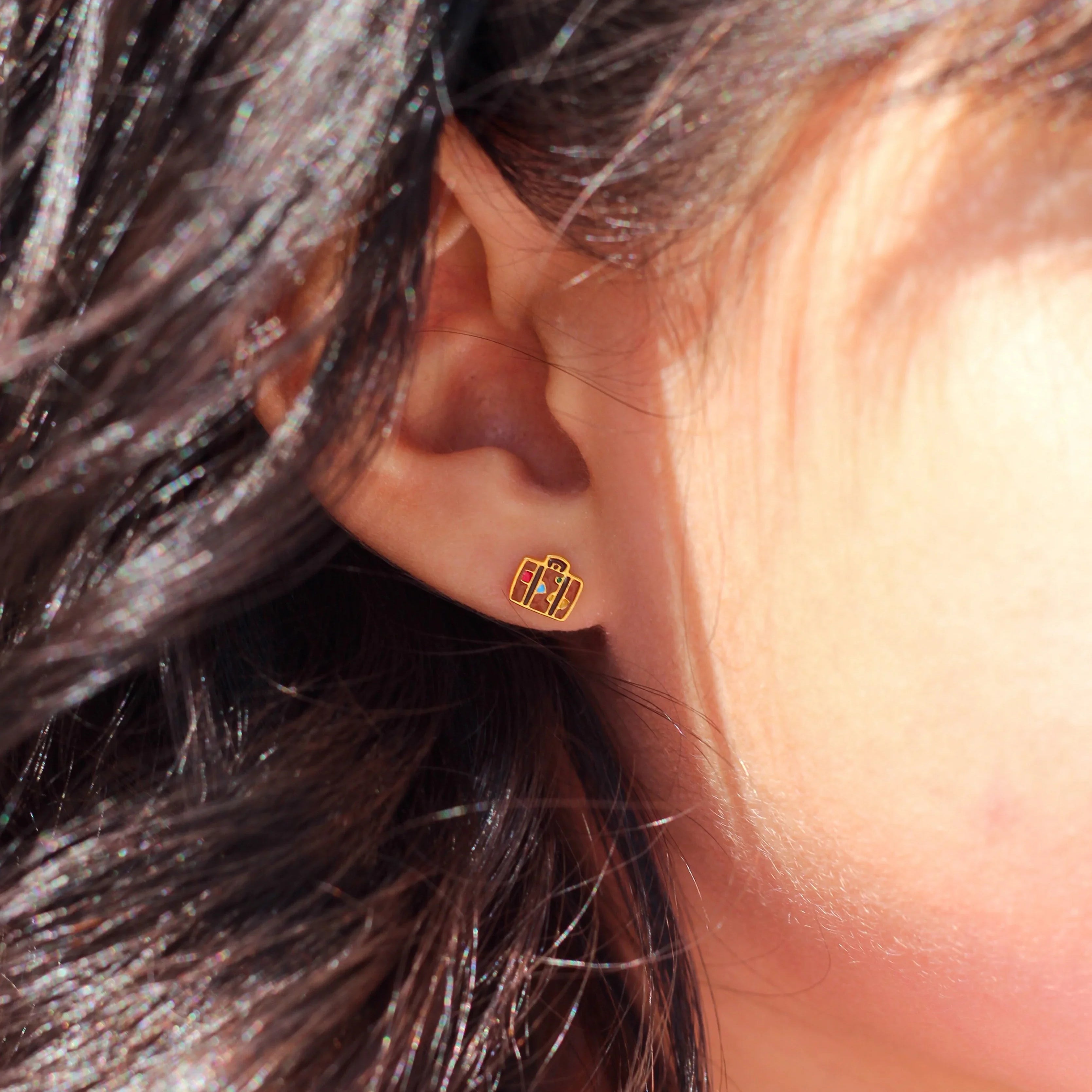 Close-up of an ear wearing a brown enamel suitcase-shaped earring with colorful shapes on it, surrounded by dark hair.