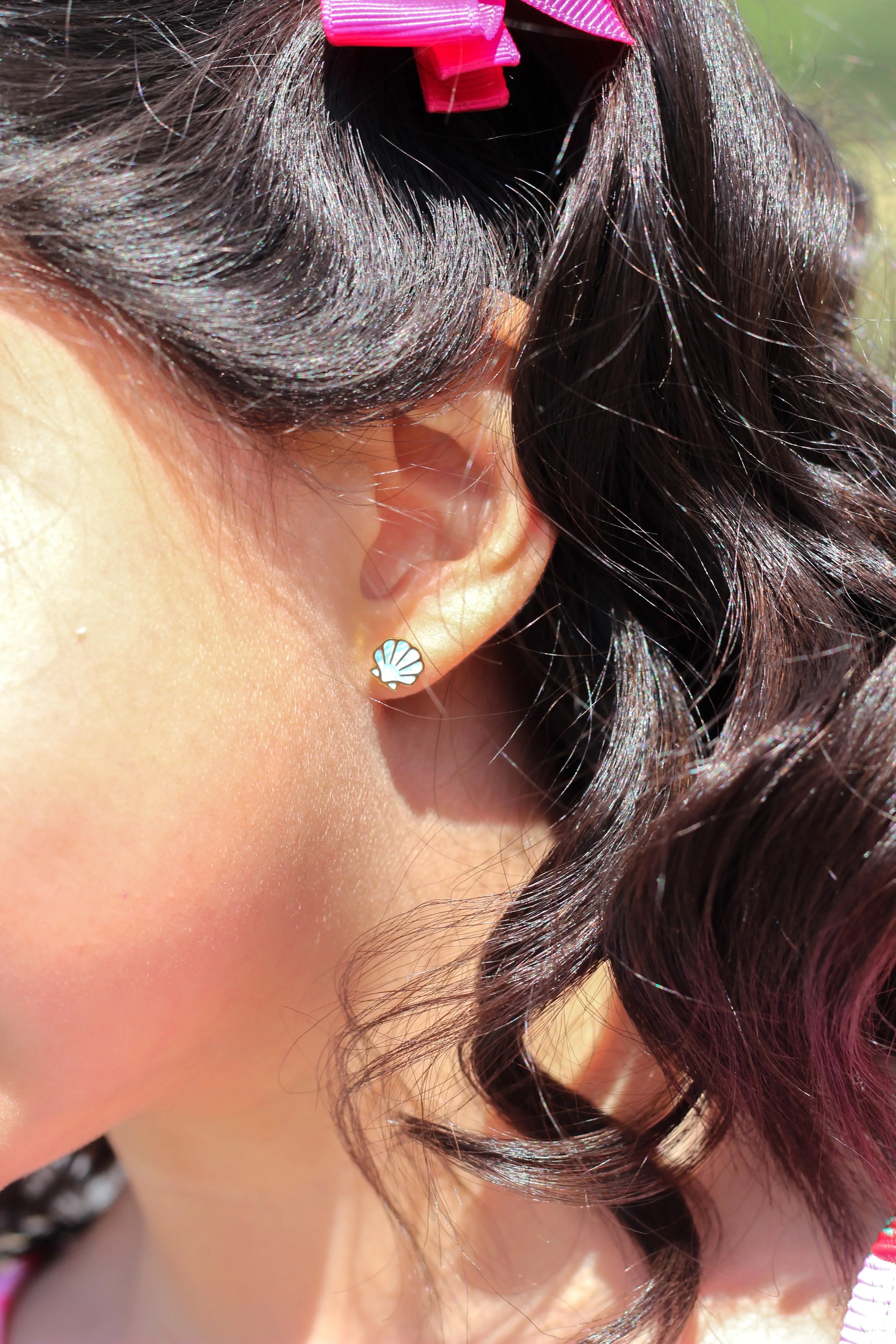 Close-up of an ear with a blue shell-shaped earring, hair with a pink clip, and blurred outdoor background.