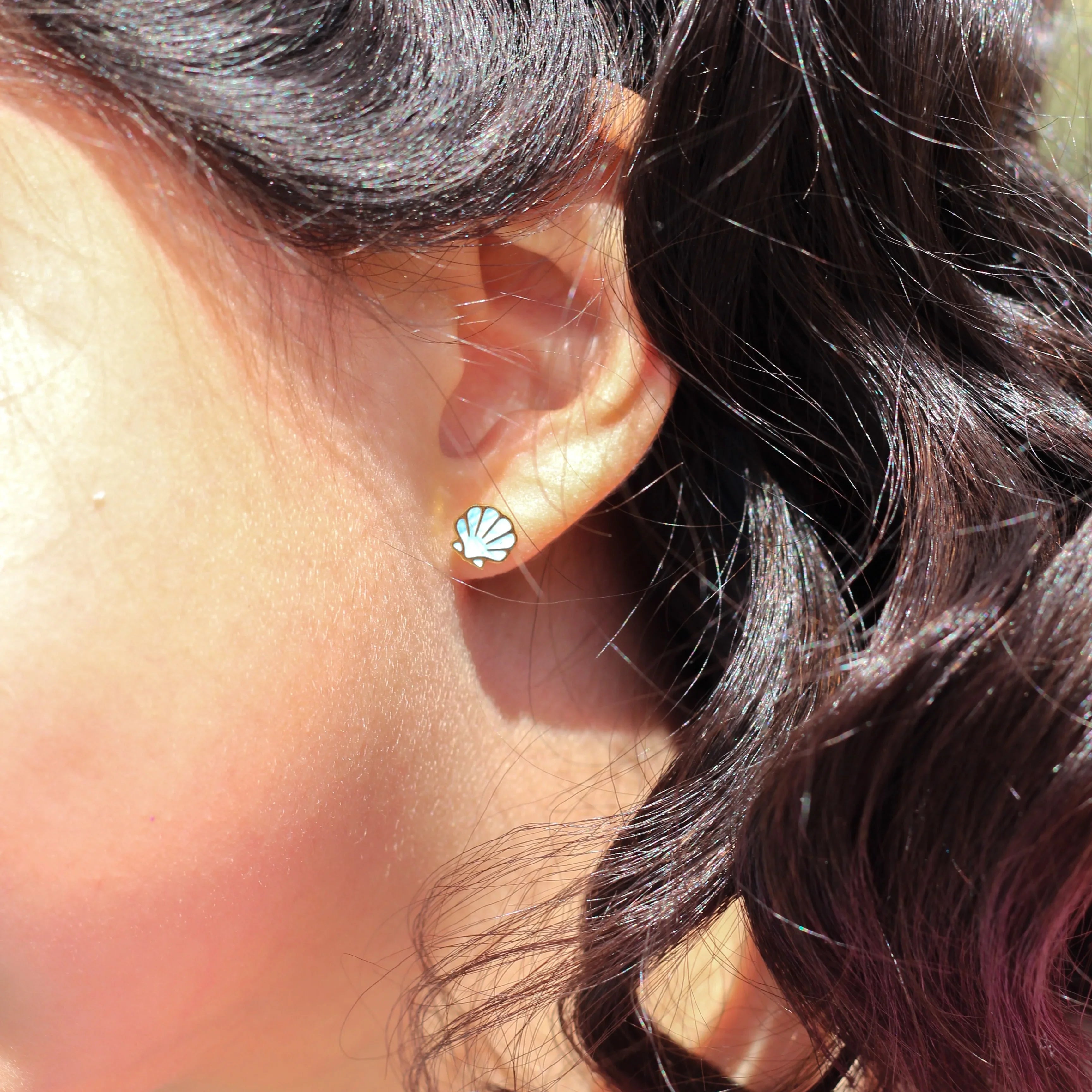 Close-up of an ear with a blue shell-shaped earring, hair with a pink clip, and blurred outdoor background.