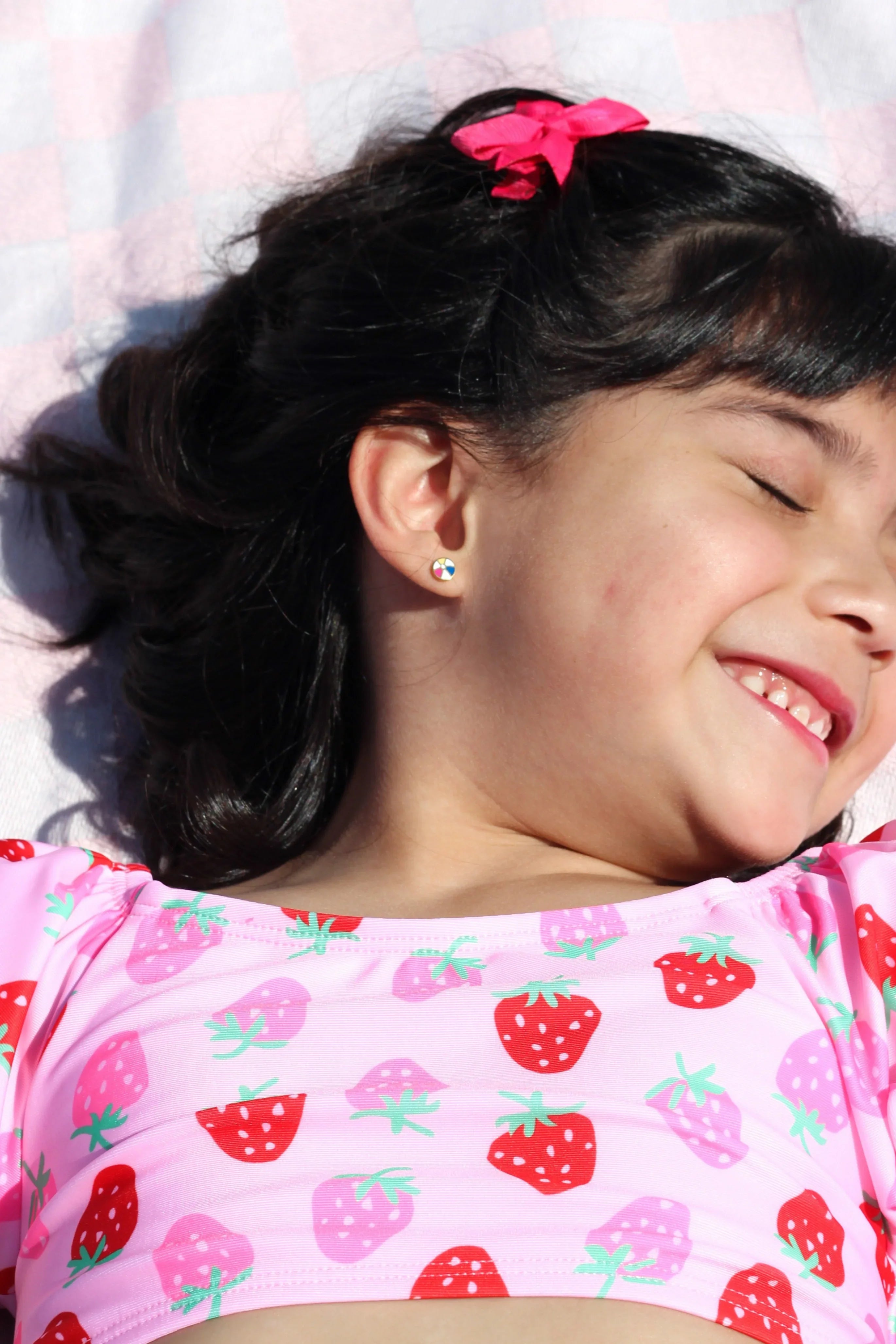 Child wearing a beach ball-shaped stud earring and a pink strawberry-patterned shirt lying on a pink checkered towel.