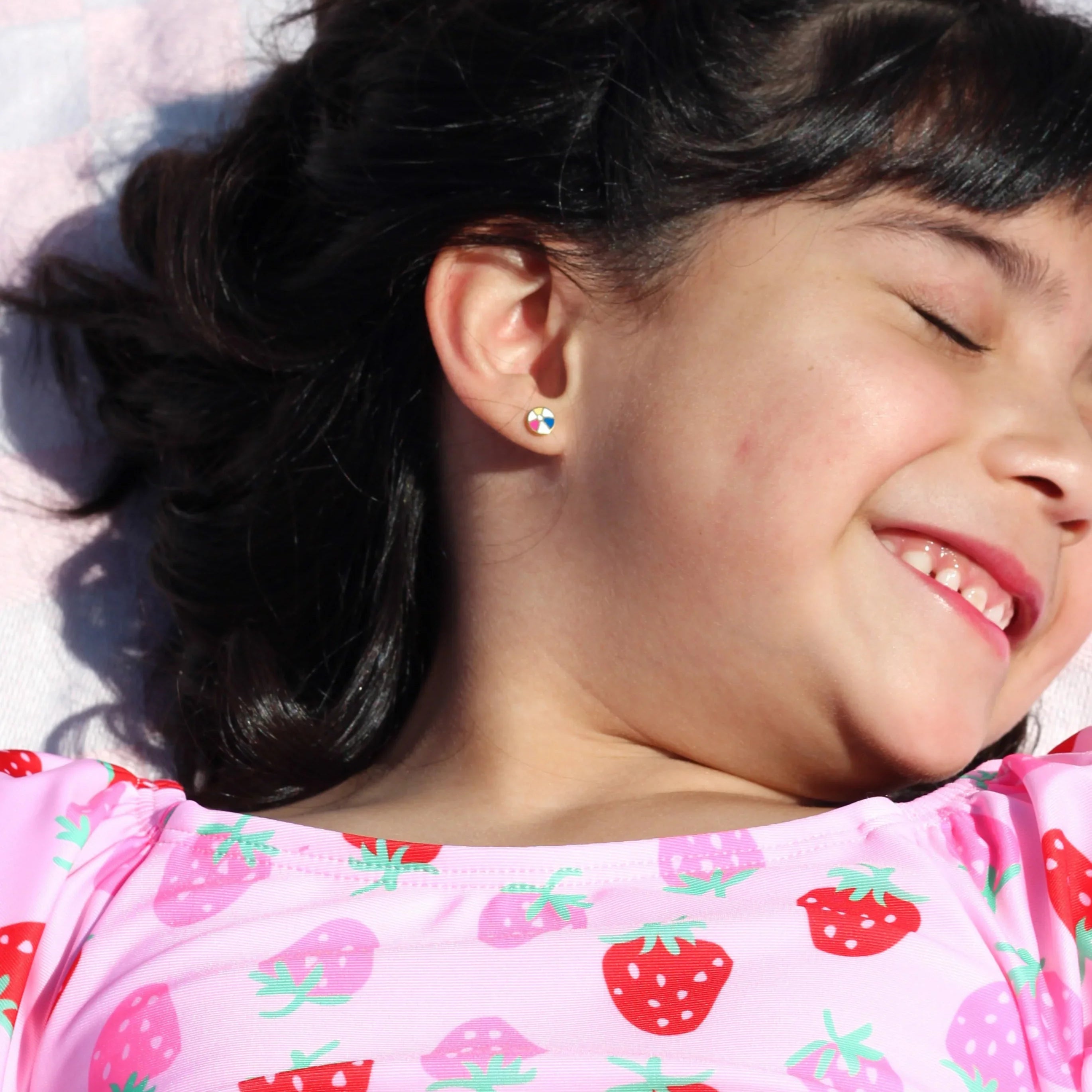 Child wearing a beach ball-shaped stud earring and a pink strawberry-patterned shirt lying on a pink checkered towel.