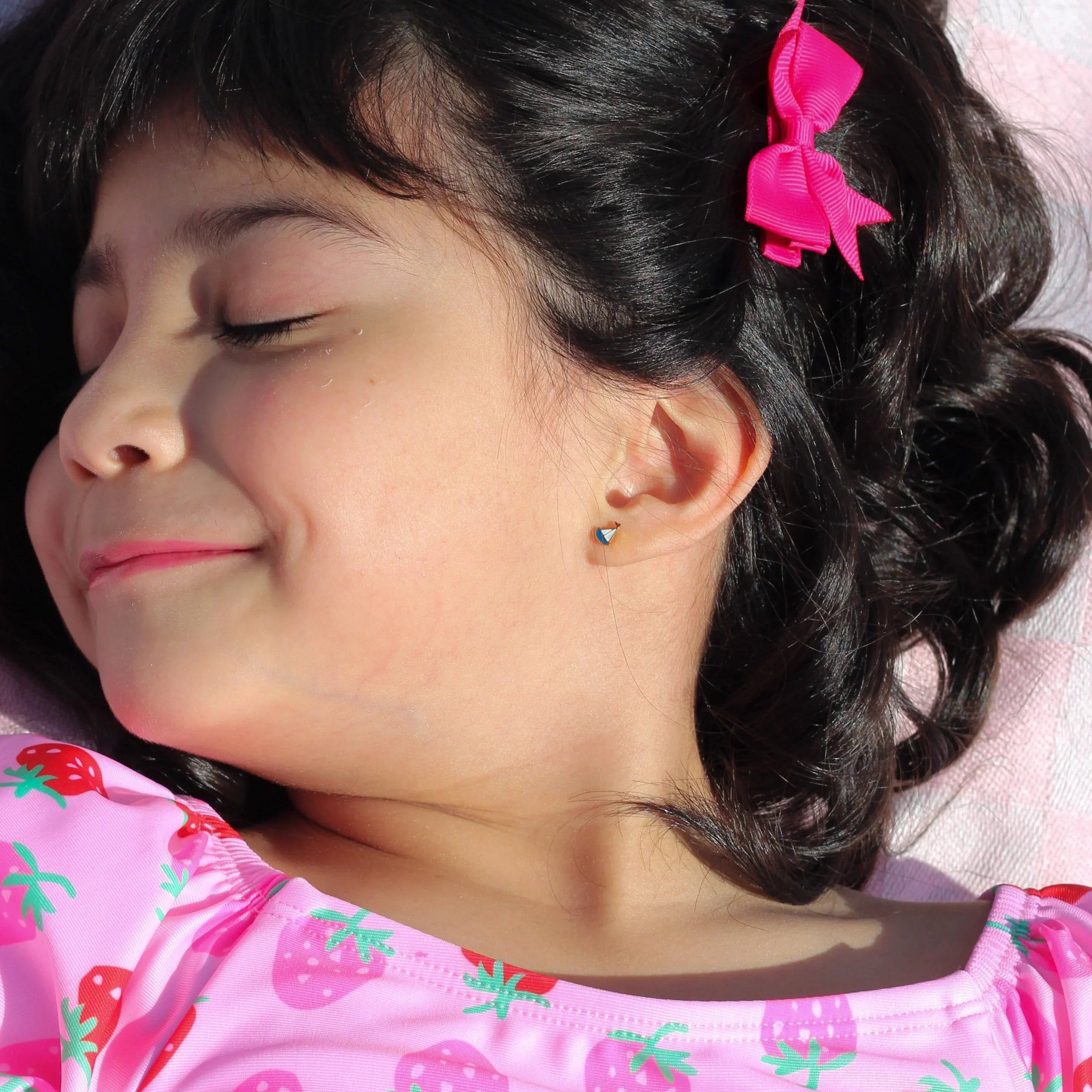 Young girl with a pink bow in her hair, wearing a pink shirt with strawberry pattern, lying on a checkered blanket.