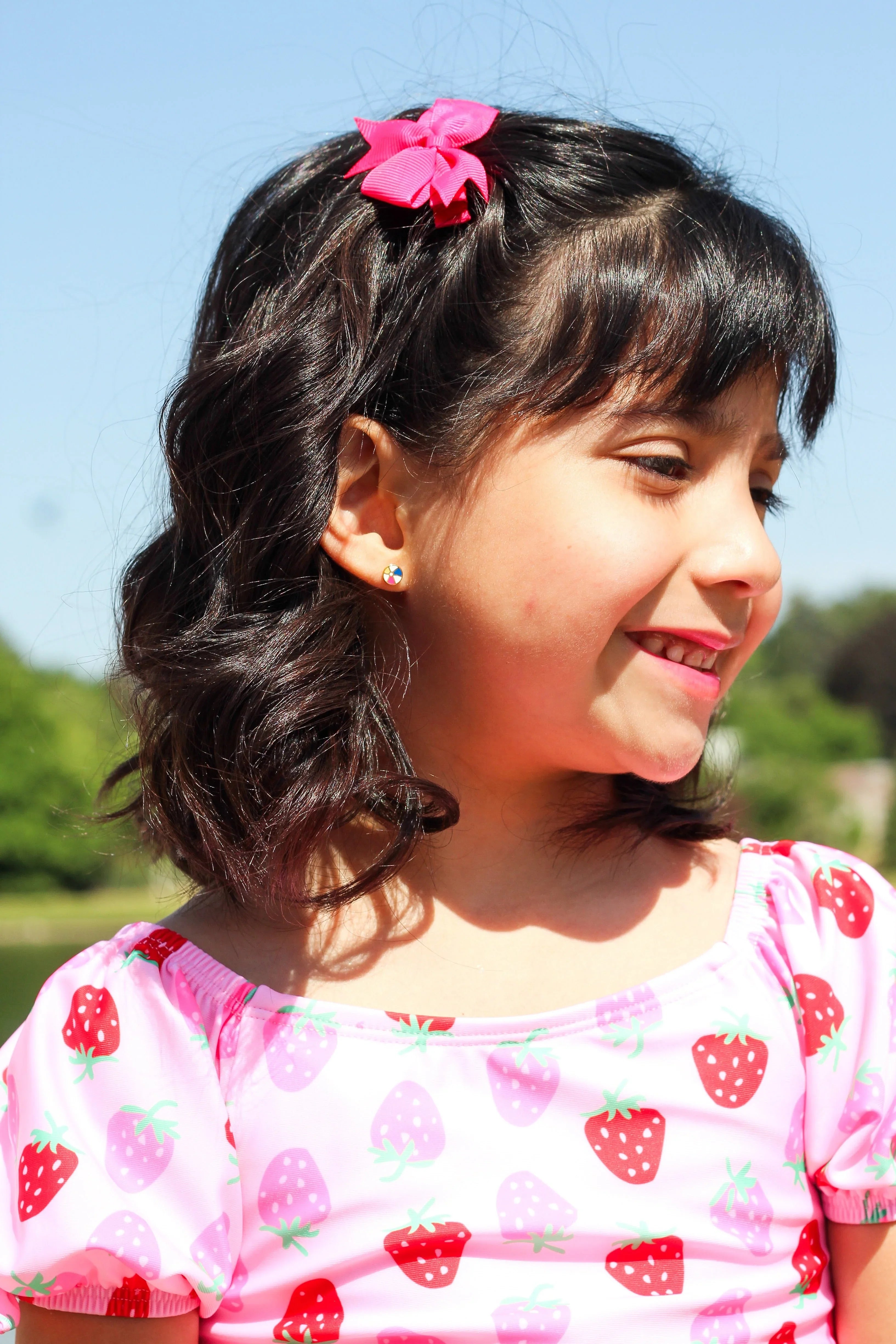 Girl with a pink hair clip, a beach ball shaped earring, wearing a strawberry-patterned shirt, outdoors on a sunny day.