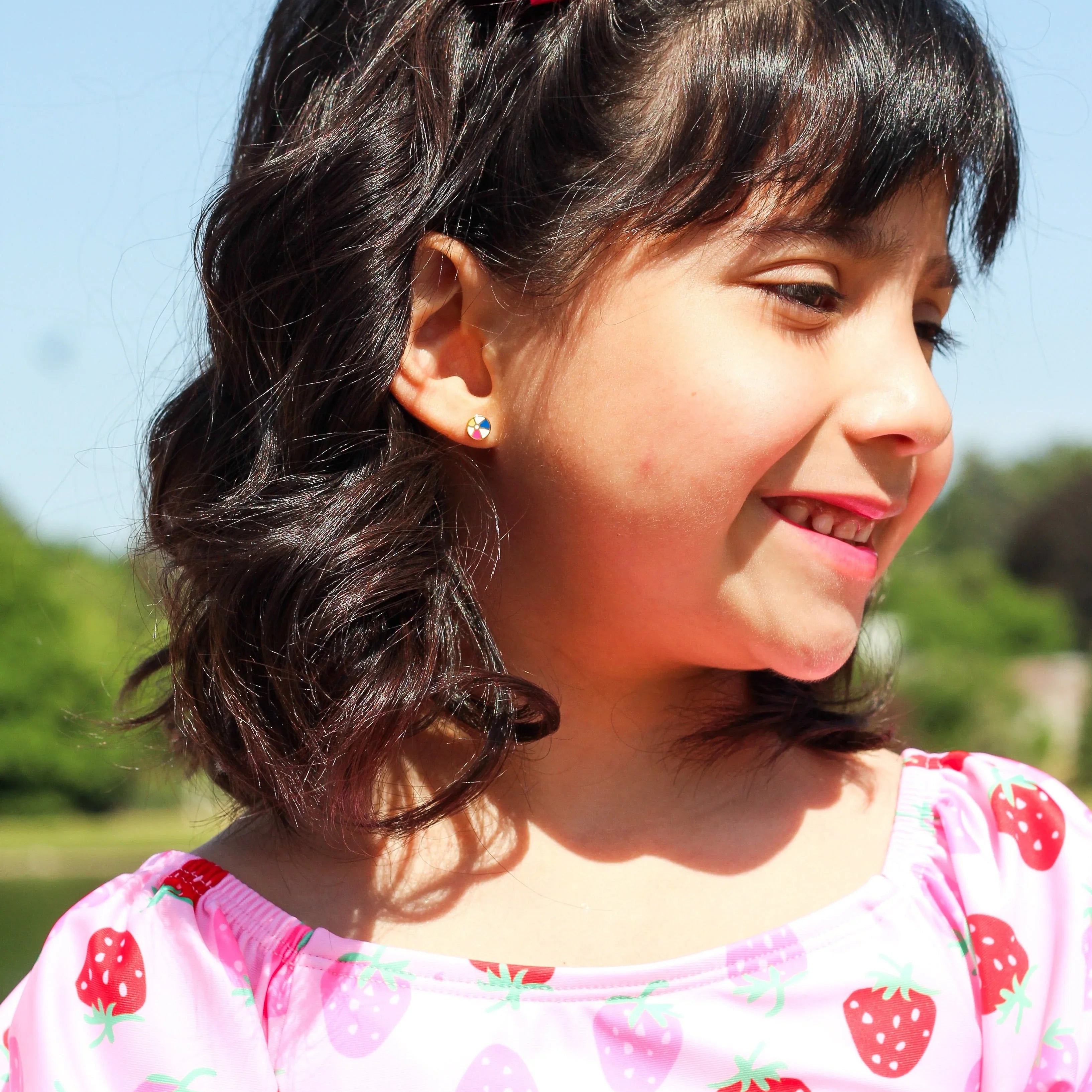 Girl with a pink hair clip, a beach ball shaped earring, wearing a strawberry-patterned shirt, outdoors on a sunny day.