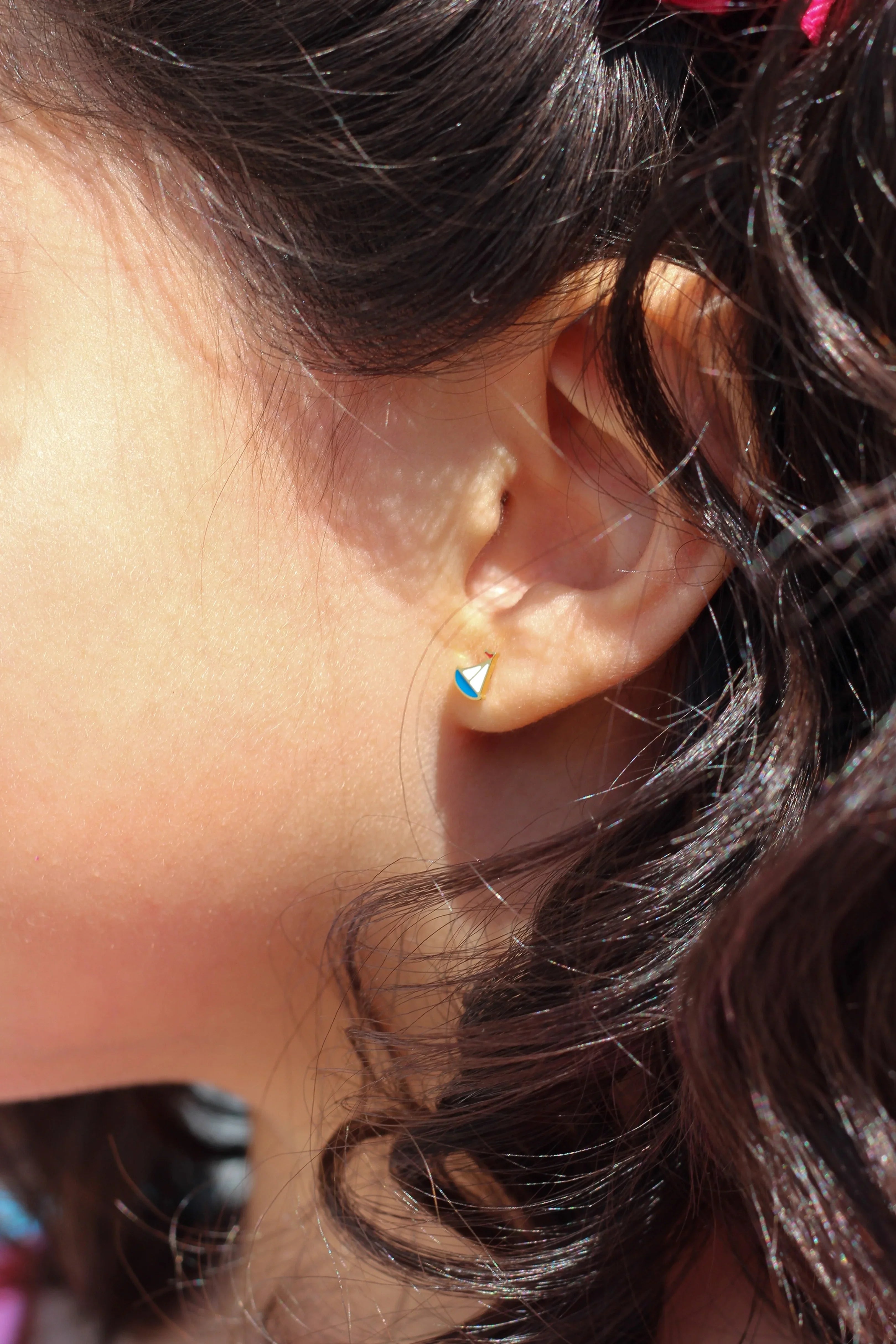Close-up of an ear with a small sailboat earring, hair tied back with a pink bow.