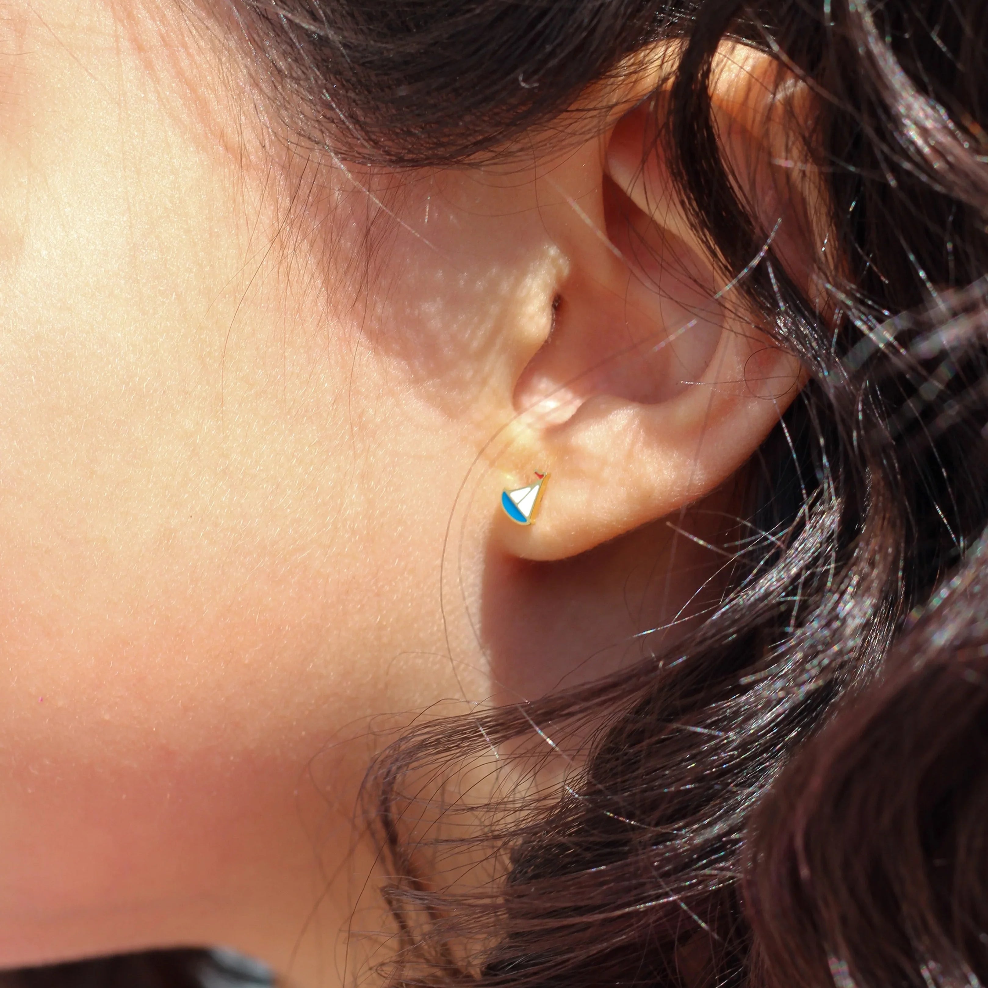 Close-up of an ear with a small sailboat earring, hair tied back with a pink bow.