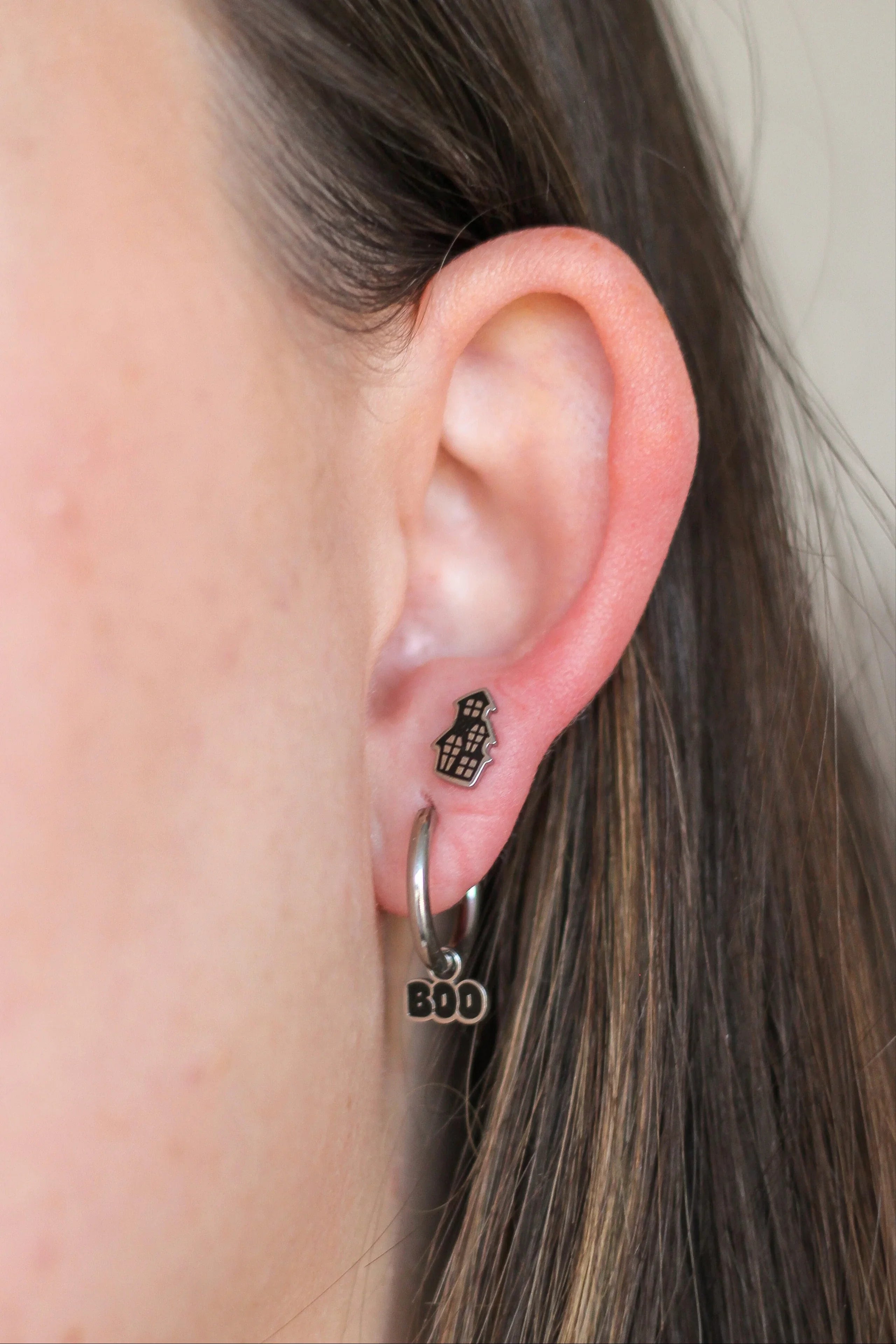Close-up of an ear with hoop earrings and a small stud that is shaped like a black and silver haunted house.